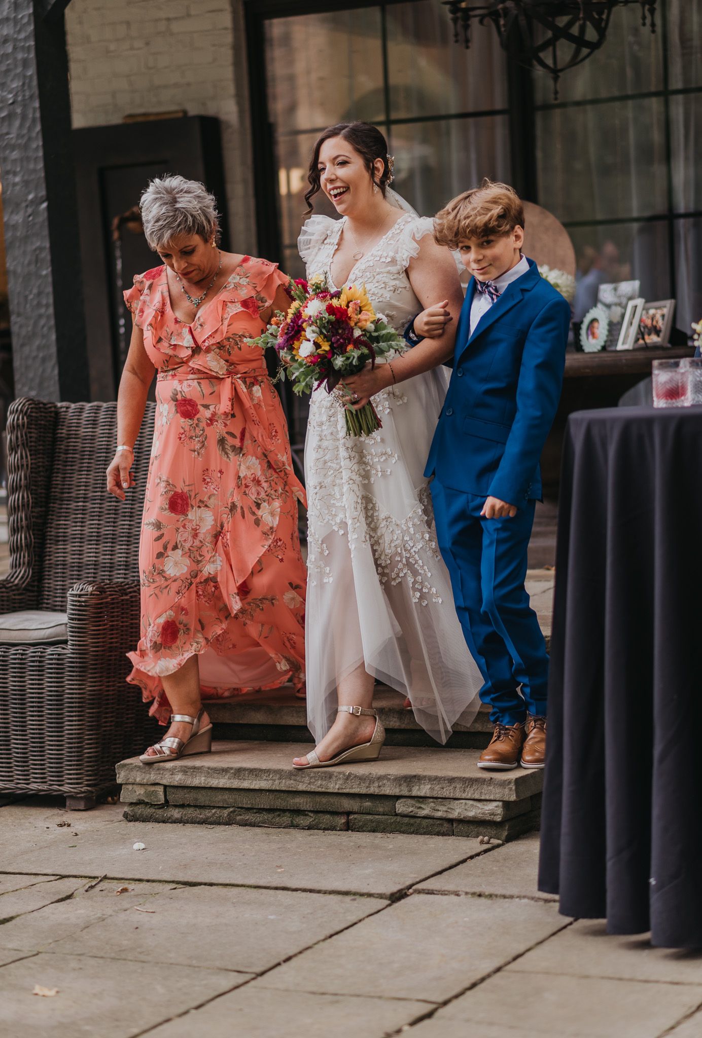 Wedding party in coral dresses and blue suits sitting on outdoor patio furniture.