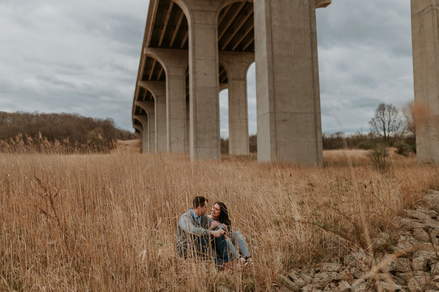 A couple stands together under a concrete bridge structure in a field of tall brown grass on an overcast day.