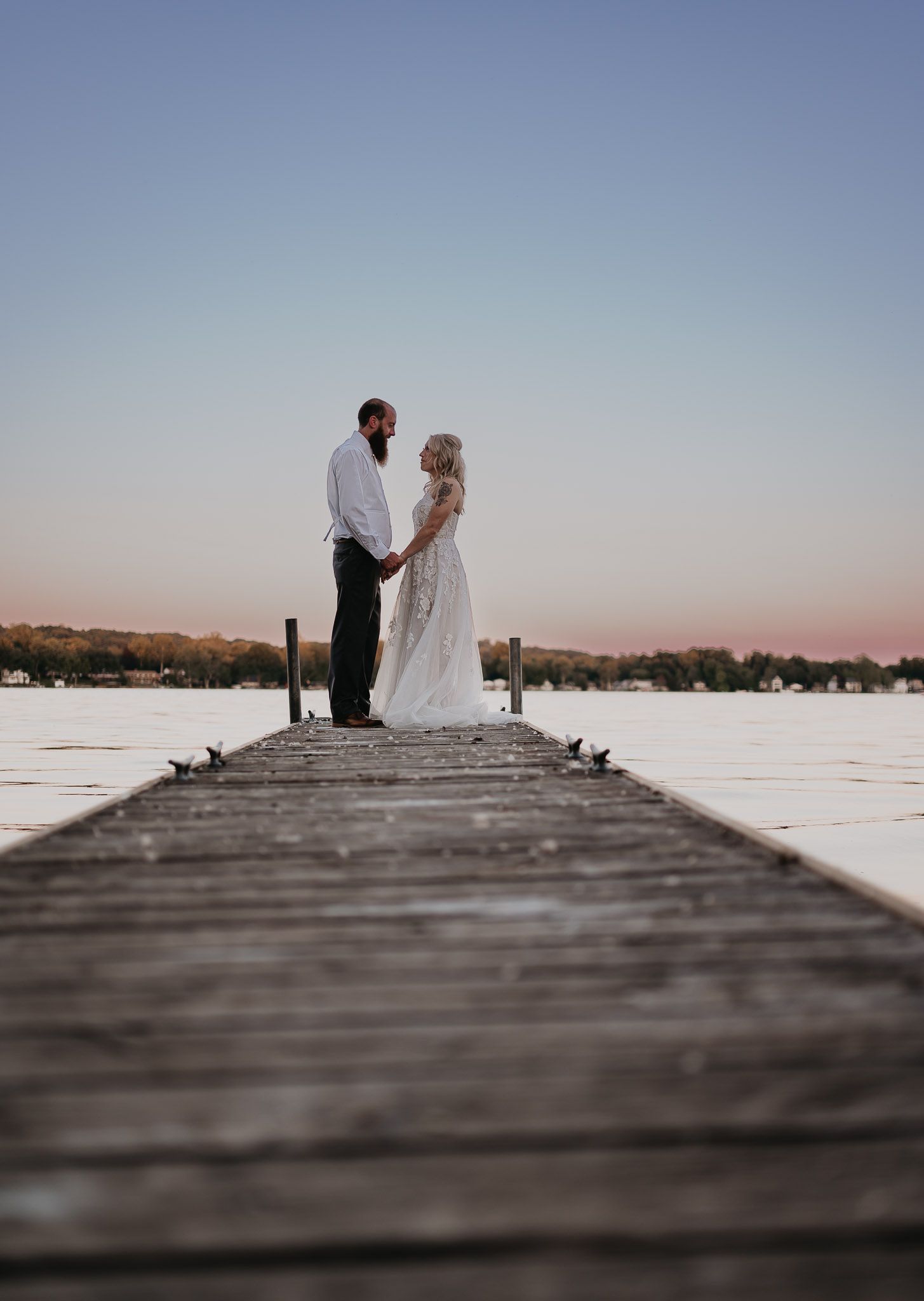 A romantic couple stands at the end of a wooden pier during golden hour.
