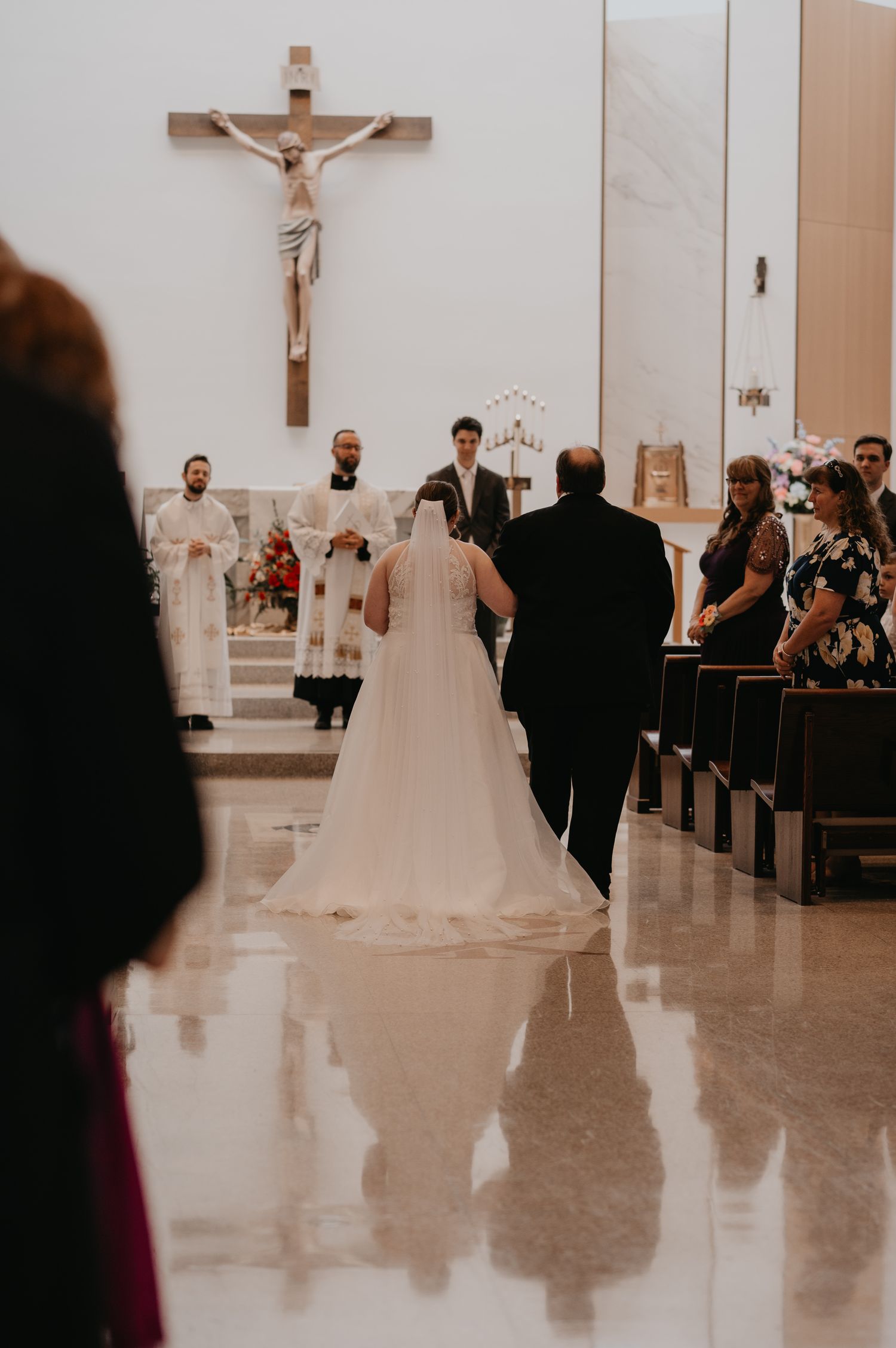 A couple walks down the aisle of a church with a crucifix visible on the wall during their wedding ceremony.