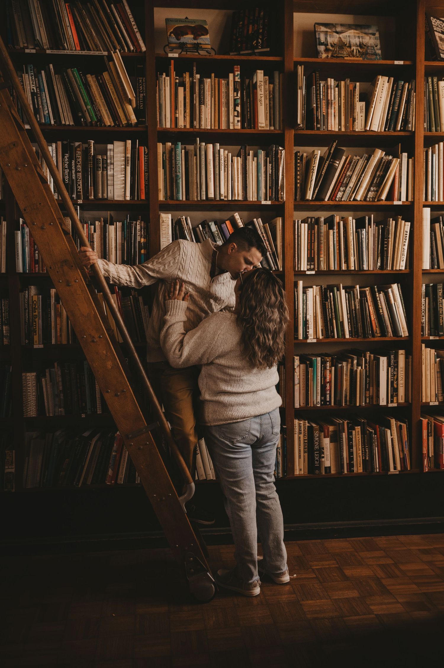 Person climbing wooden ladder to reach books on tall shelves in a dimly lit library with floor-to-ceiling bookshelves.