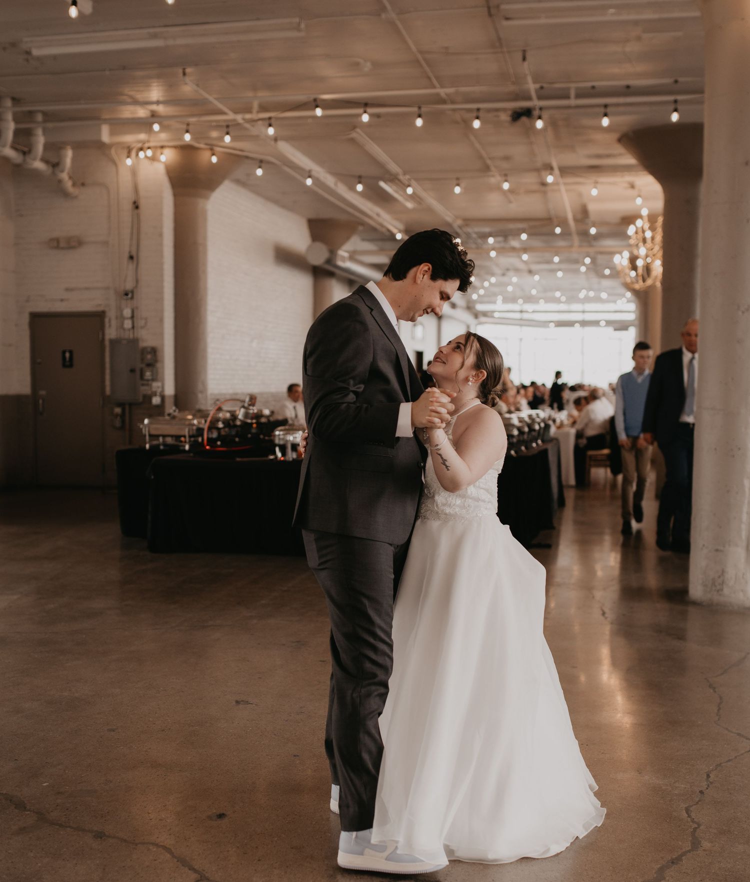 A couple shares their first dance in an industrial-style reception venue with string lights overhead.