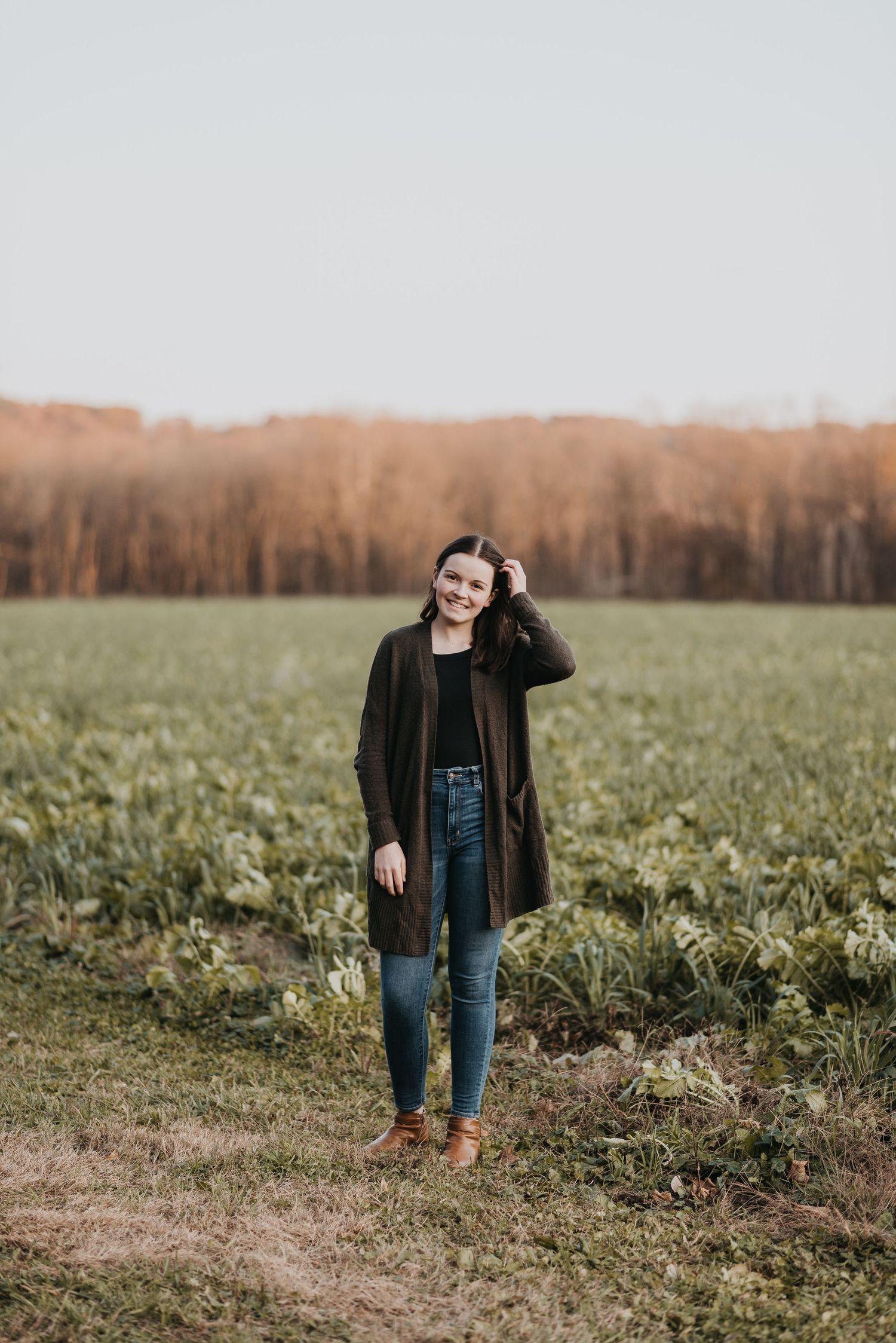 Someone stands in an open green field wearing a long green cardigan, blue jeans and brown boots at sunset.