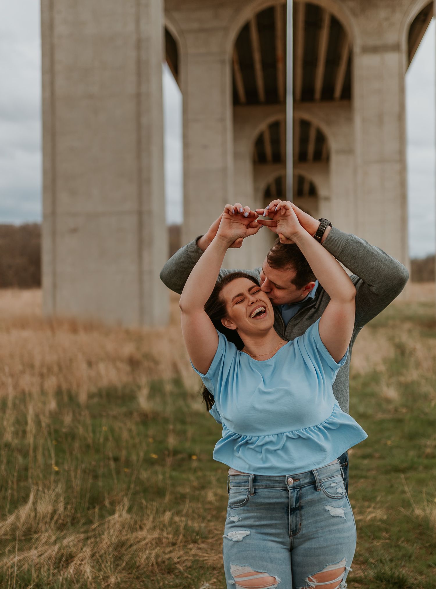 A person laughs while making heart gestures with their hands beneath a concrete overpass.