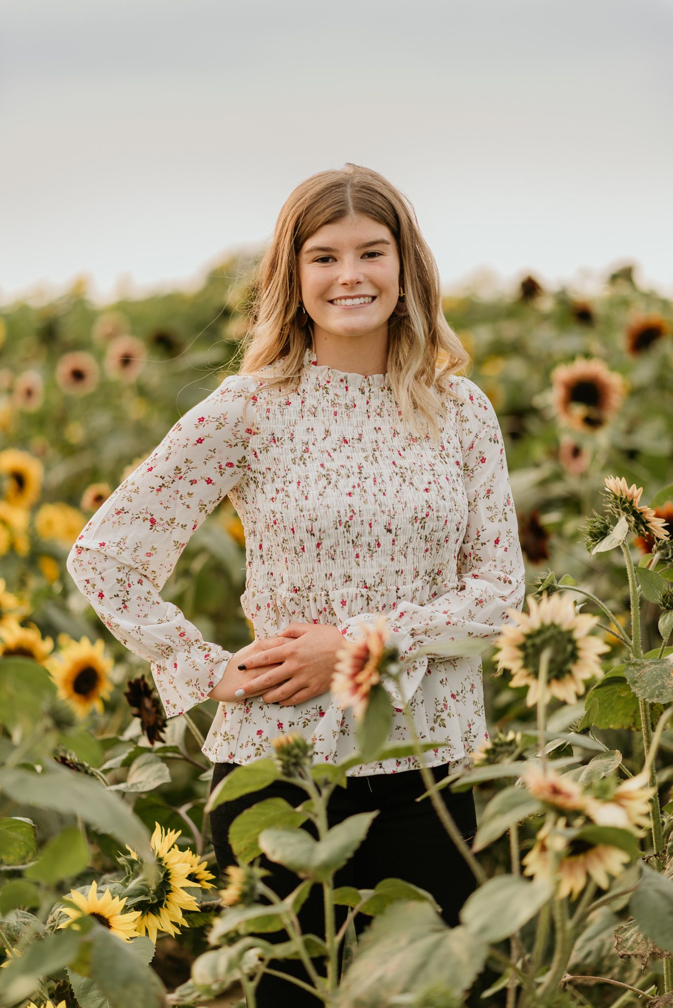 A person with blonde hair wearing a white floral top poses surrounded by vibrant blooming sunflowers in a field.
