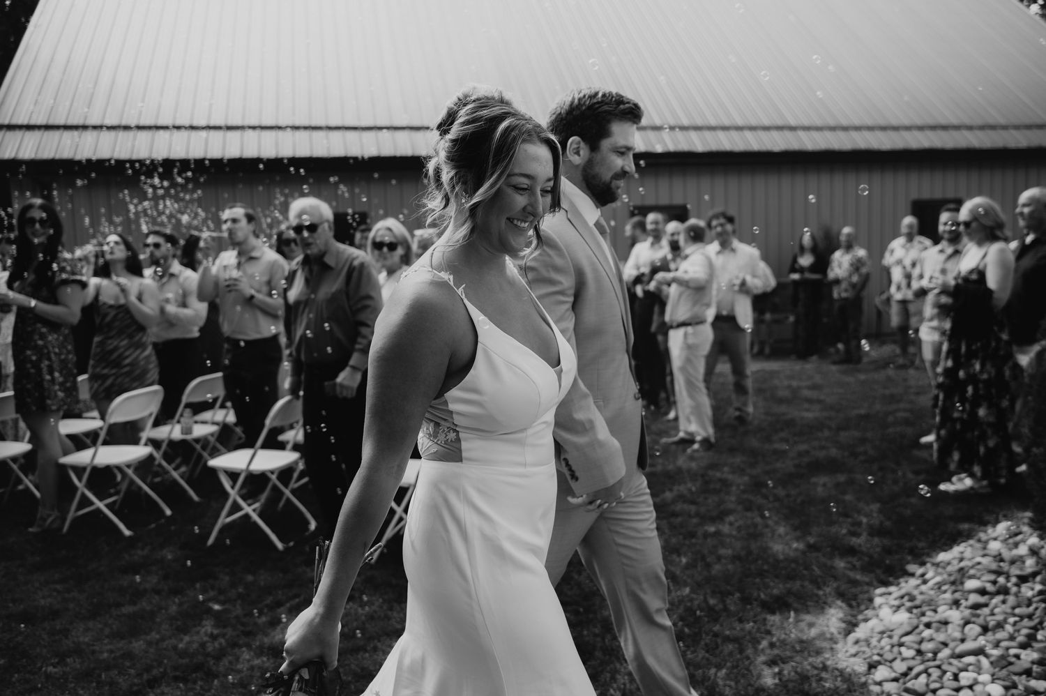 A couple walks together at their outdoor wedding reception while guests look on in black and white photography.