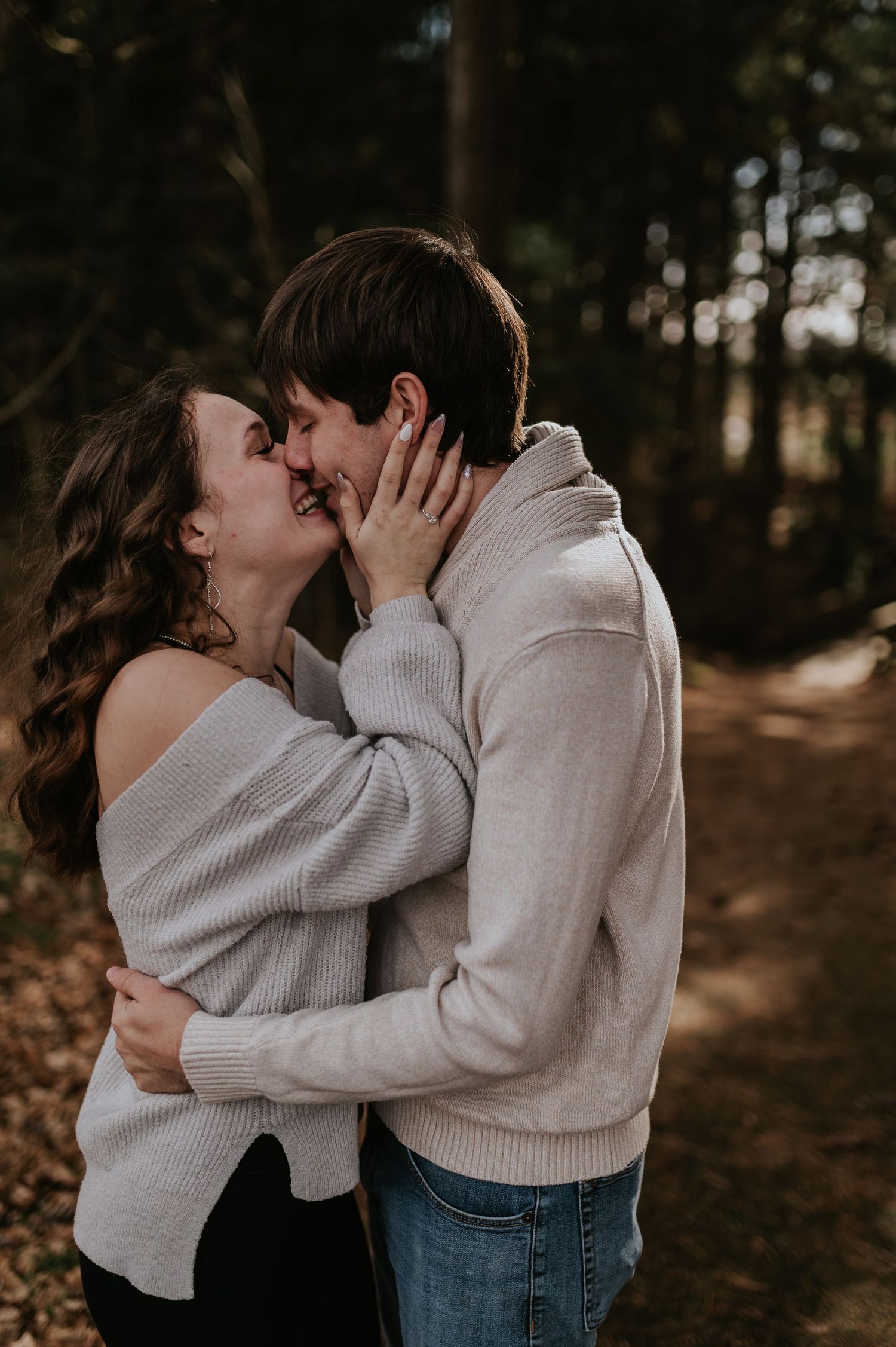 Couple in coordinated cream sweaters sharing a romantic moment on a forest path.