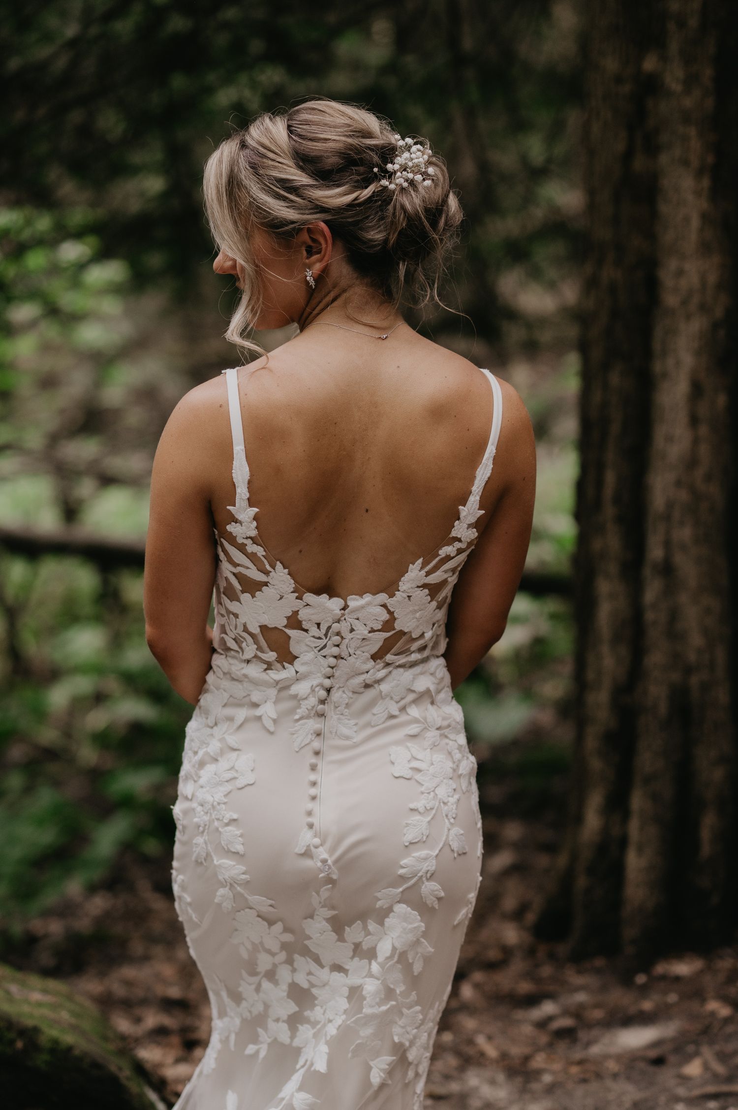 Back view of a delicate lace wedding dress with intricate floral details against a forest backdrop.
