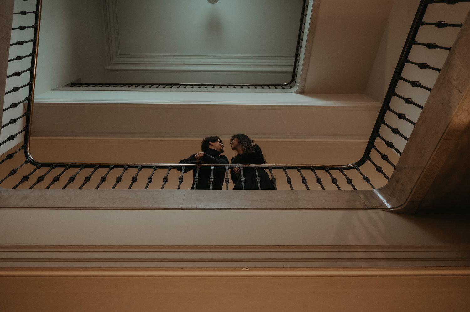 Looking down a spiral staircase with white painted walls and wooden railings, capturing two figures in silhouette below.