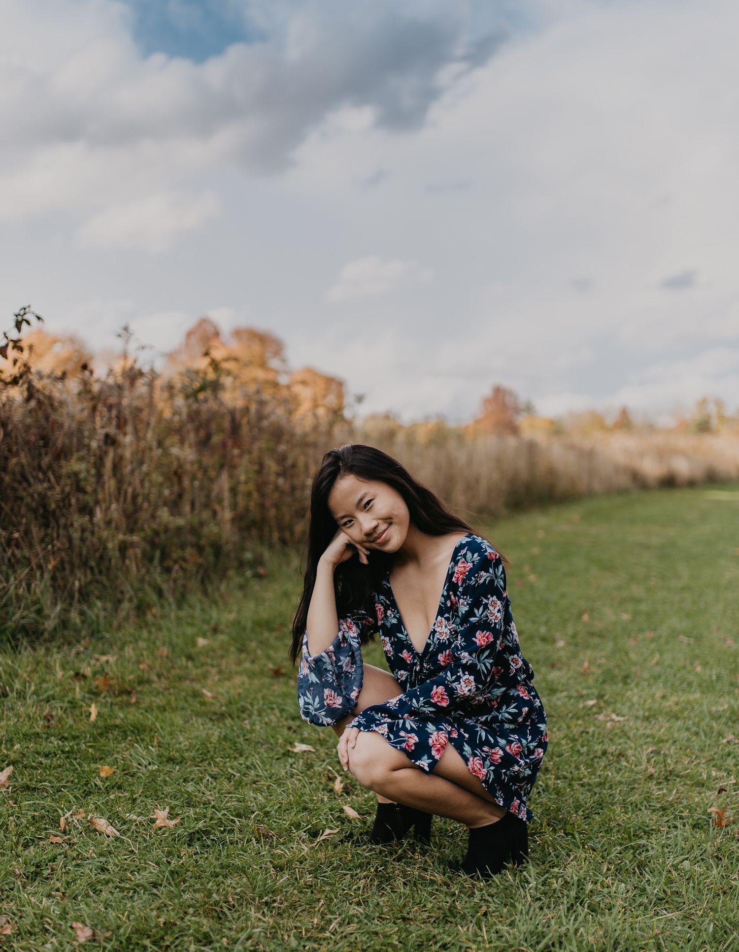 Person in navy floral dress crouches on grassy path with warm autumn colors in background.