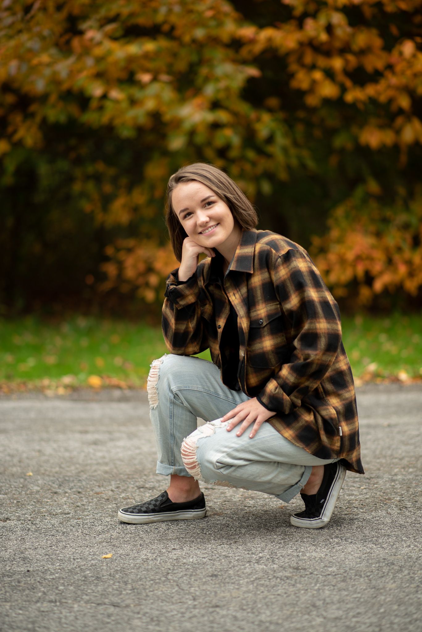 Portrait of someone crouched on pavement wearing a plaid jacket and jeans against autumn foliage background.