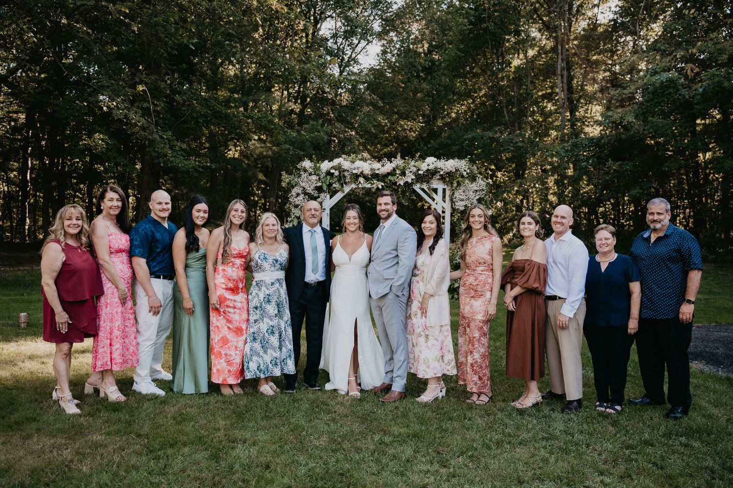 A large wedding party group poses together outdoors in coordinated pink, white and blue attire at sunset.