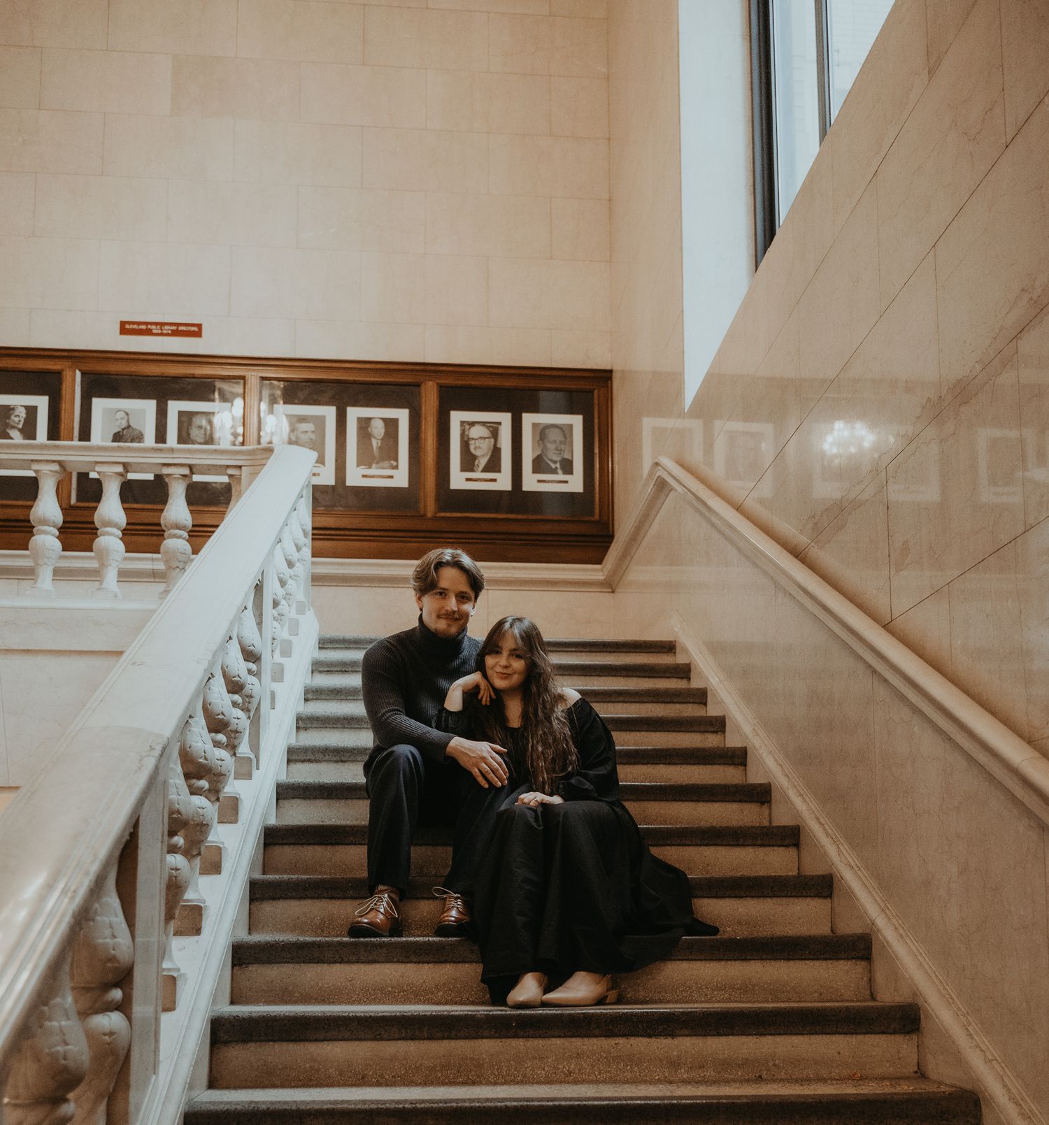 A couple sits together on stone steps in a dimly lit interior space with beige walls and framed artwork.