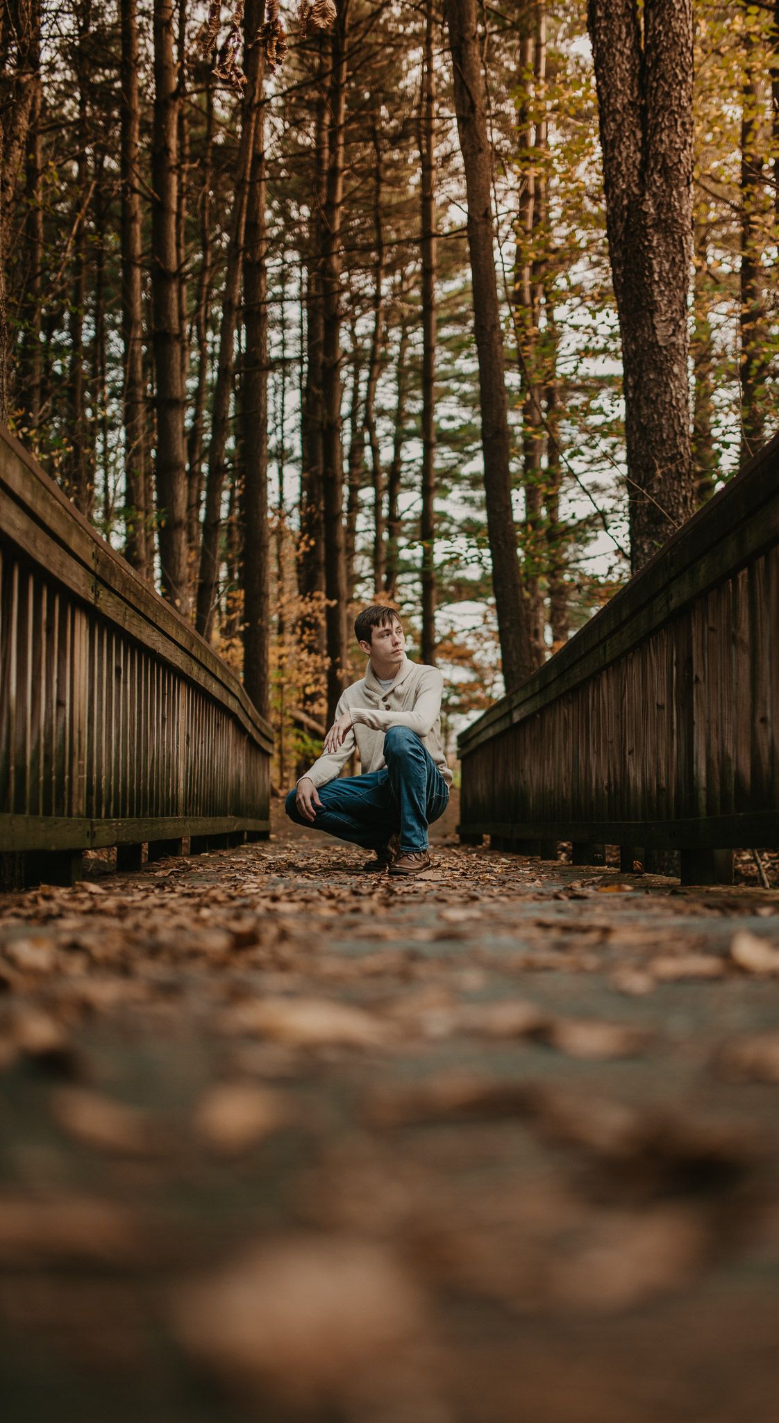 Person crouches on leaf-covered wooden boardwalk in autumn forest setting.