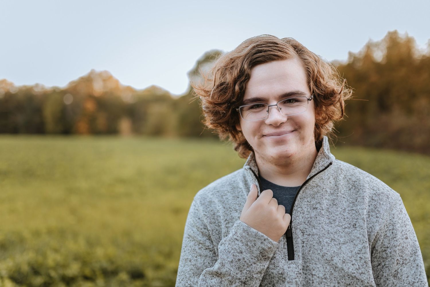 A person wearing a grey quarter-zip sweater smiles while standing in a green field during golden hour.