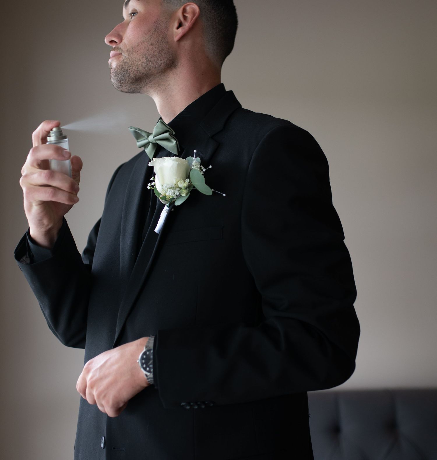 Groom in black tuxedo with green bow tie and boutonniere sprays cologne before wedding ceremony.