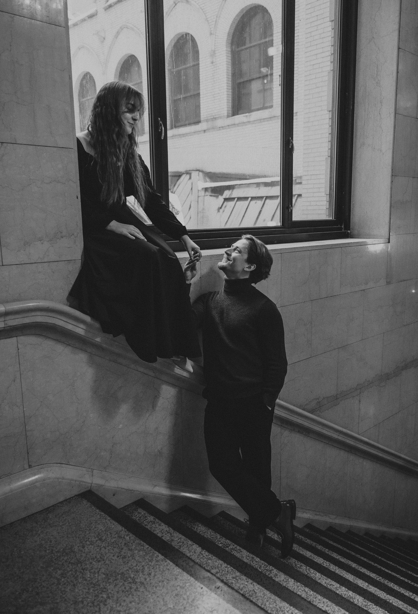 A black and white photo of two people in a stairwell with arched windows, one sitting on a ledge while the other stands below.