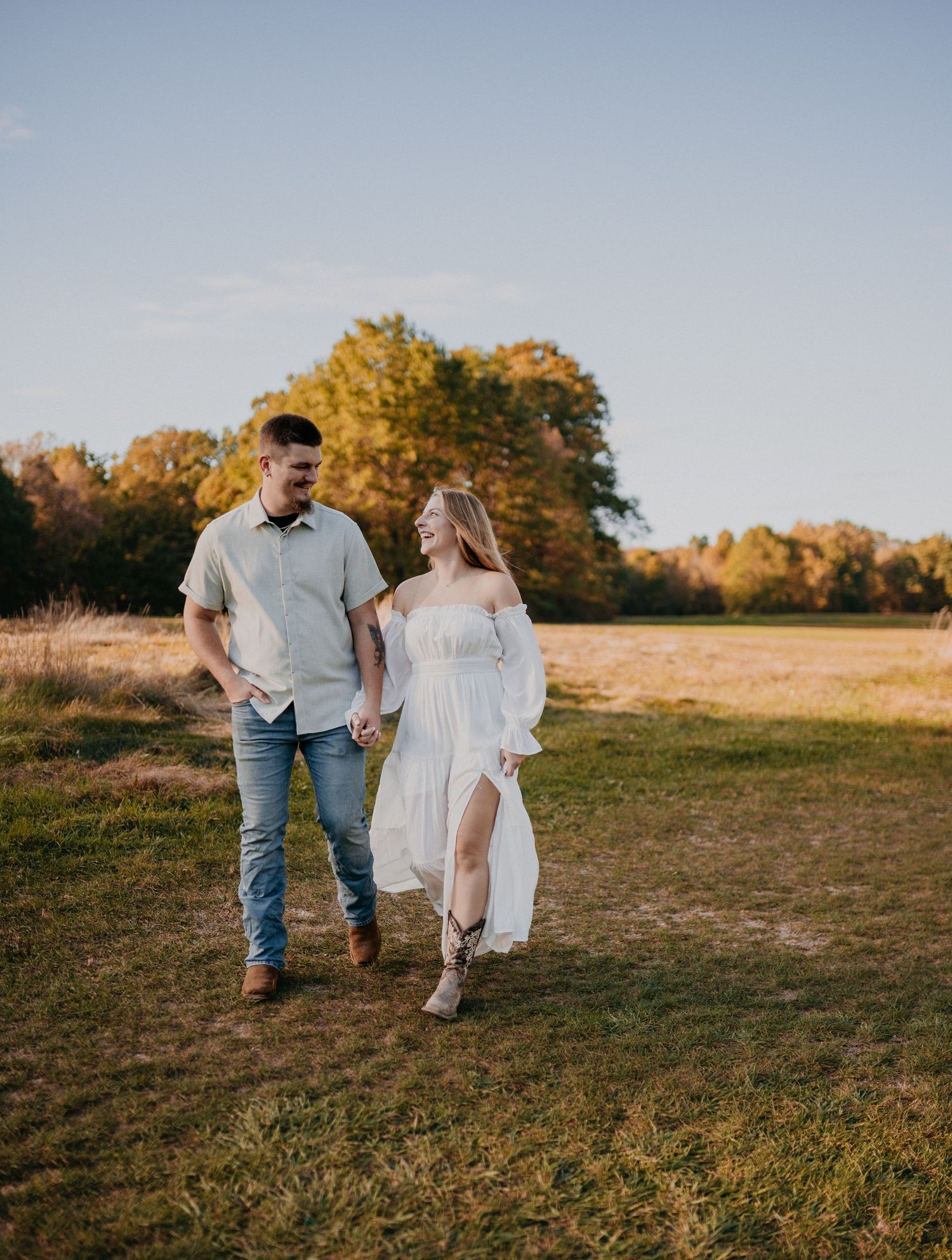 A couple walking hand in hand through a sunlit field with golden trees in the background at sunset.