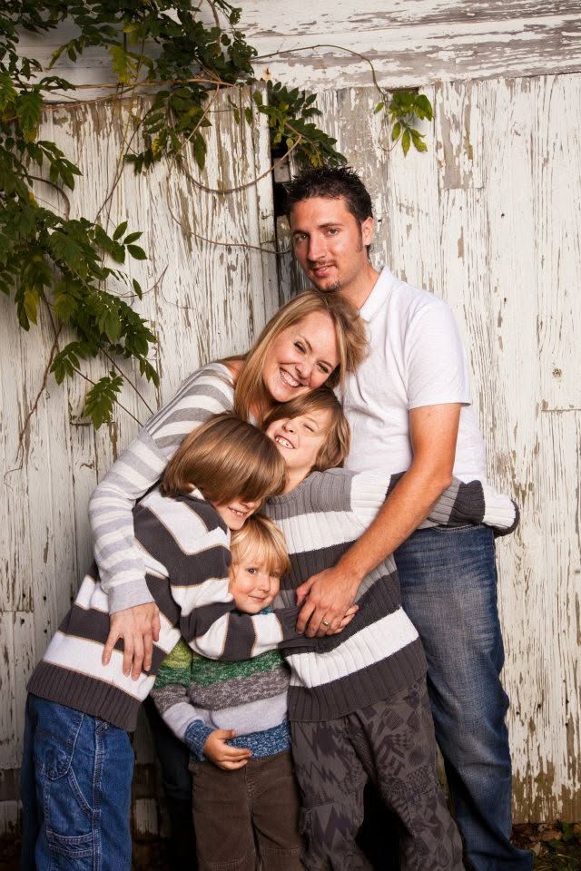 Happy family of five hugging and smiling together against a rustic white painted wooden wall with green vines.