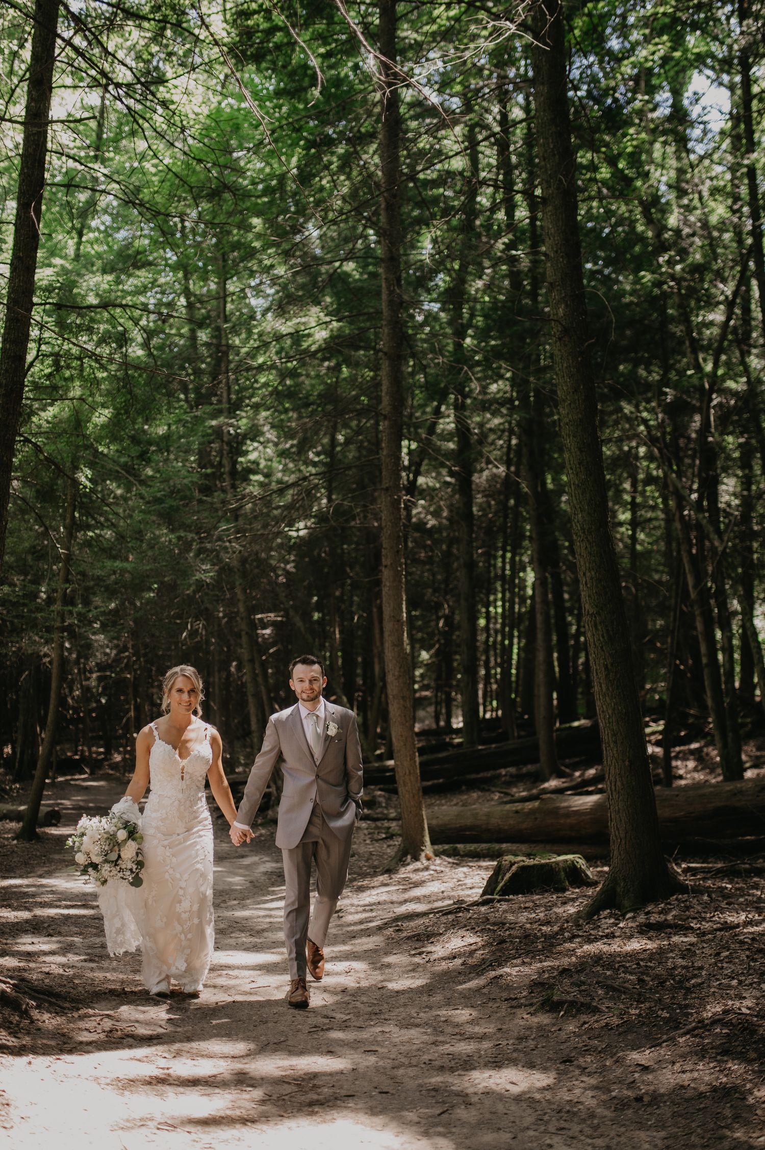 A couple in wedding attire walks hand in hand along a sunlit forest path surrounded by tall trees.