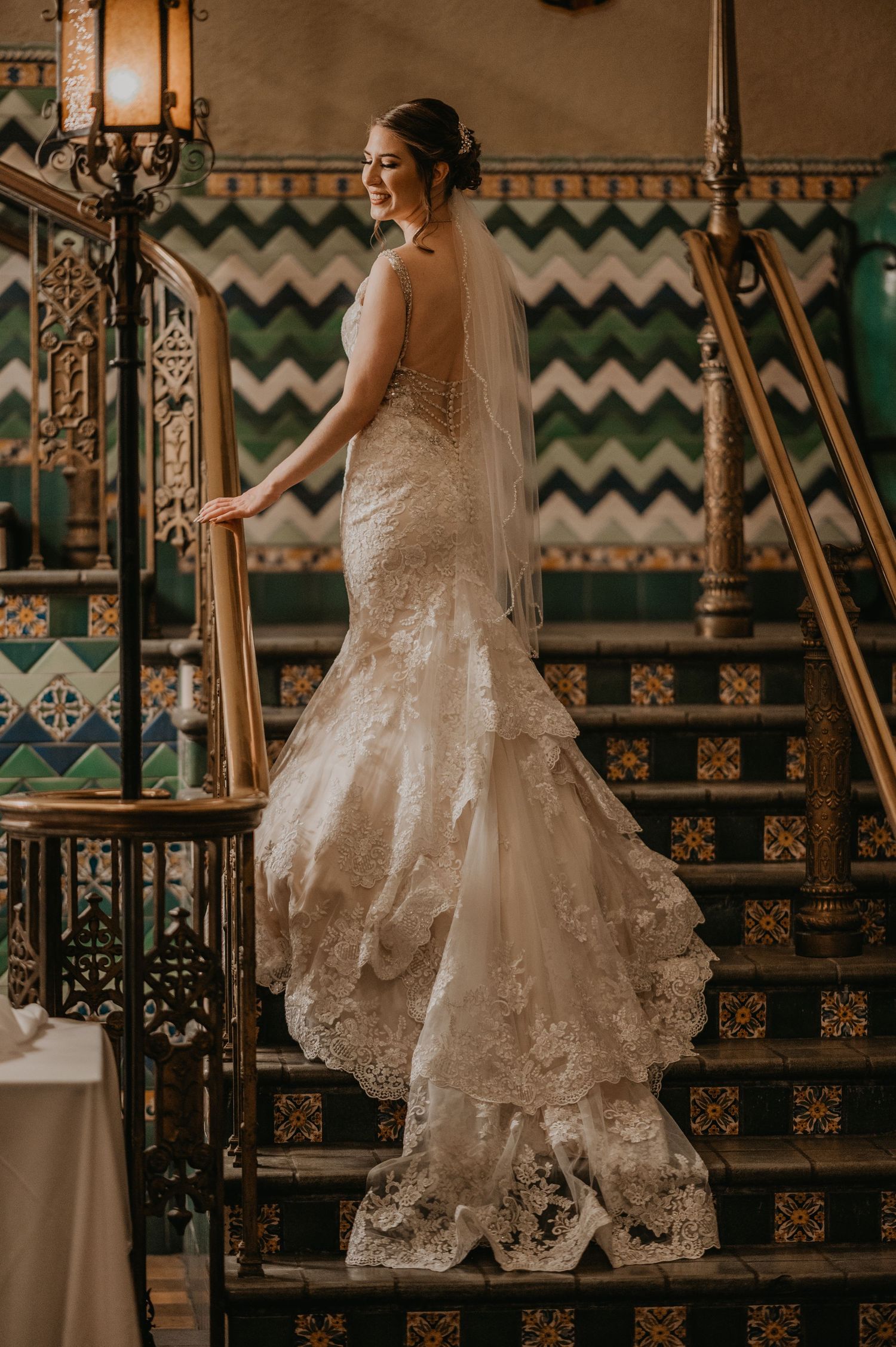 Bride in elegant lace wedding gown poses on ornate vintage staircase.