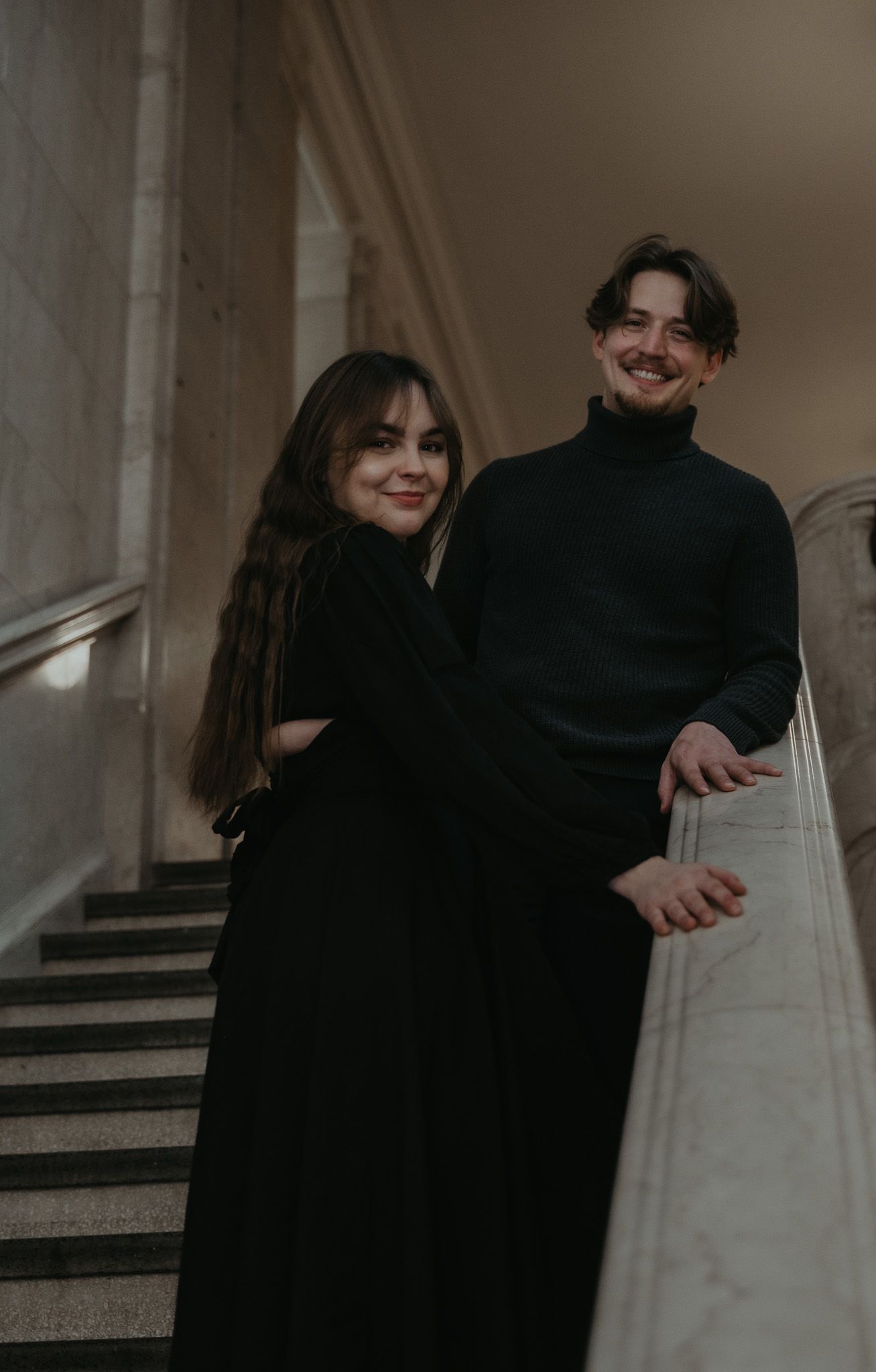 A couple in dark clothing embraces on a dimly lit staircase in a moody and romantic atmosphere.