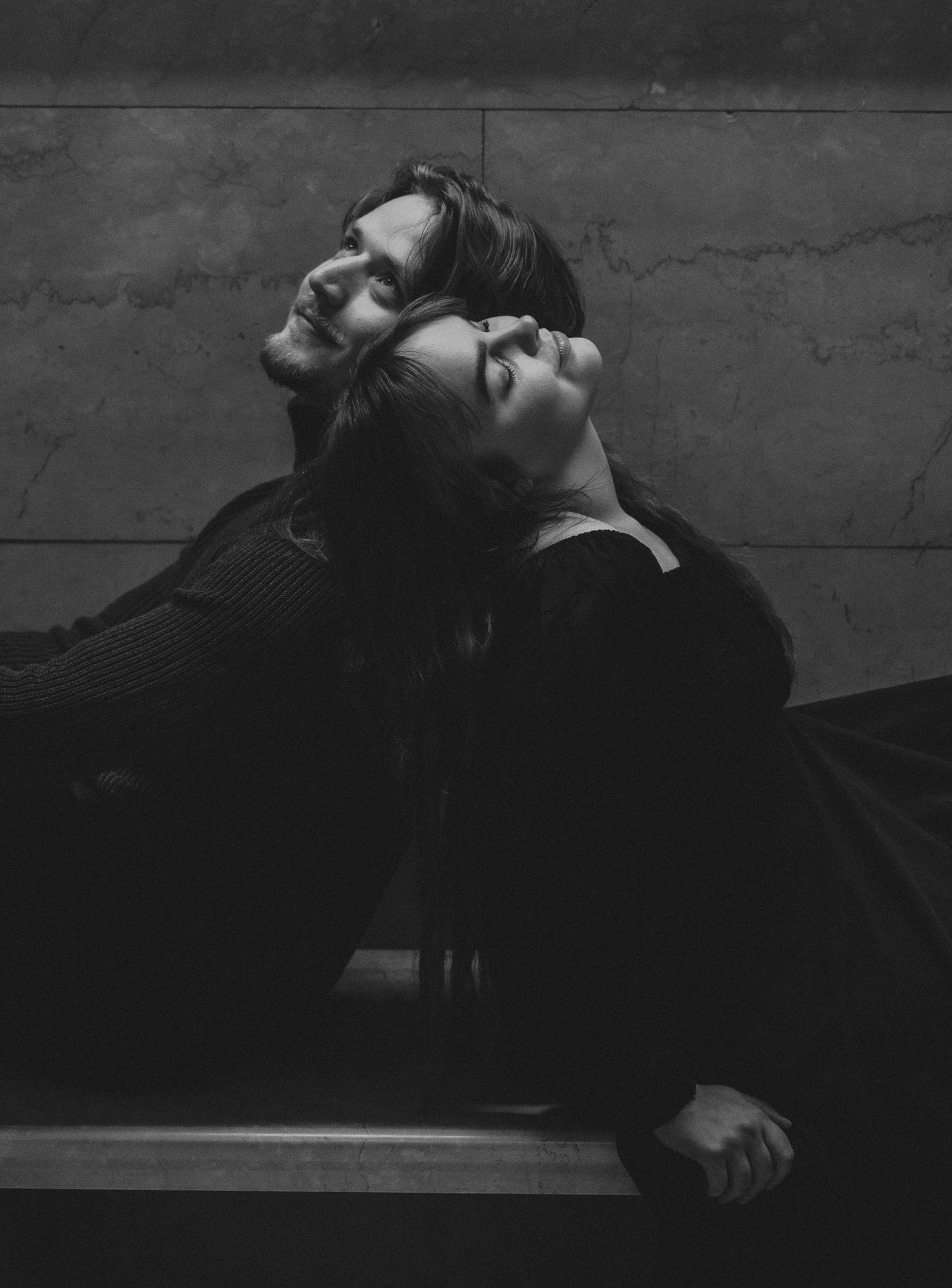 Artistic black and white portrait of two people resting their heads back to back against a textured wall.