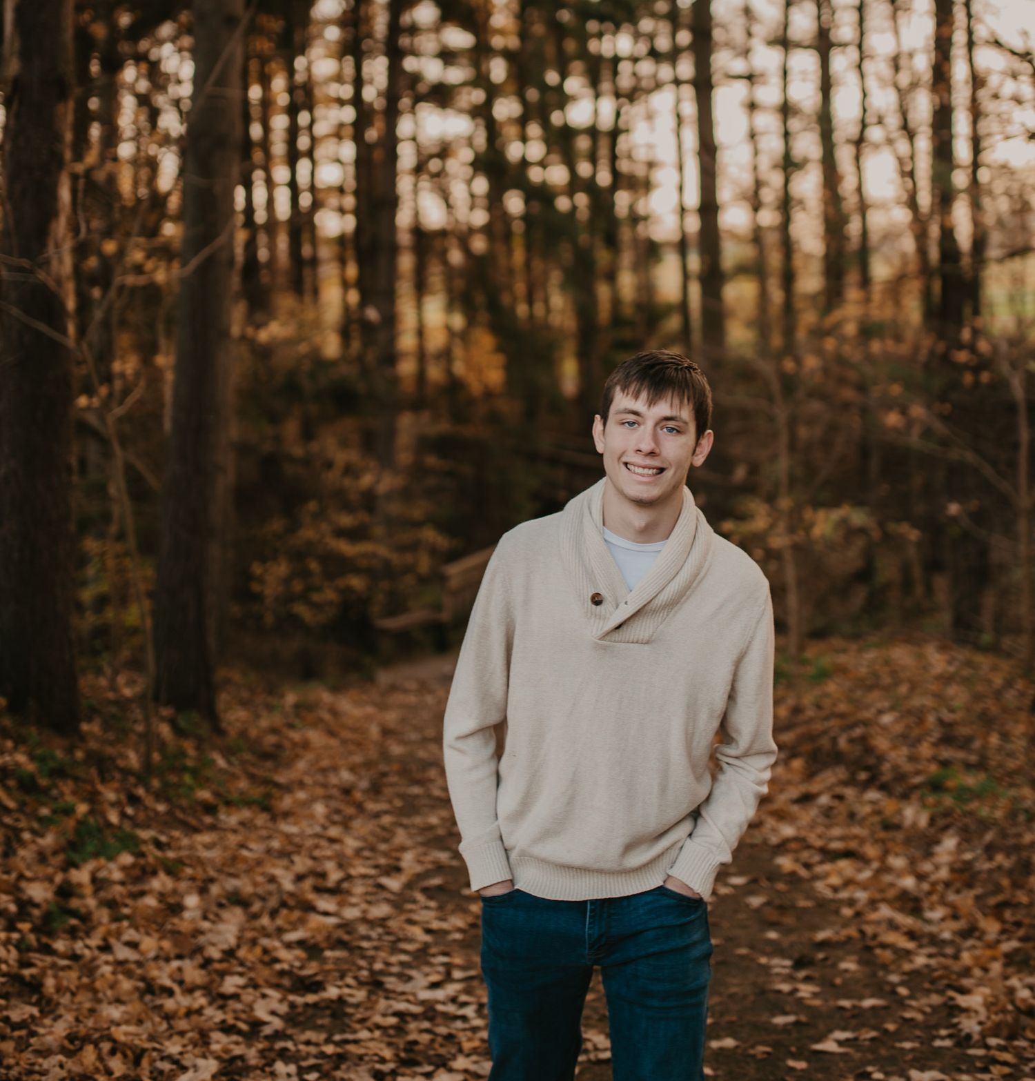 Standing on a forest path surrounded by fall foliage wearing a light-colored pullover.