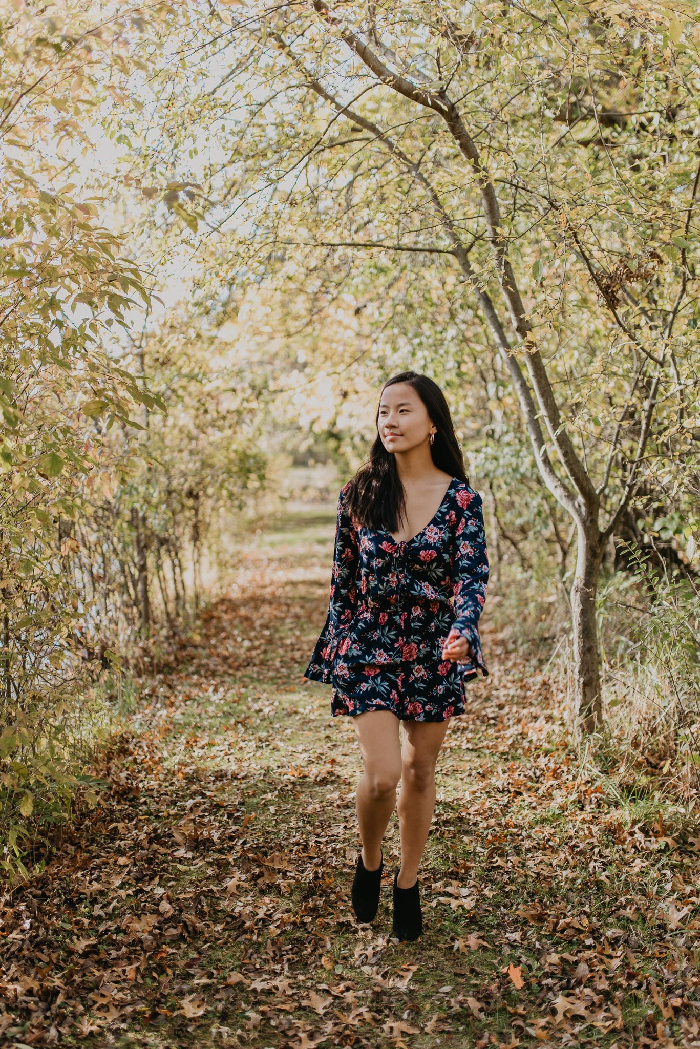 Person in floral dress walking through wooded area with fallen leaves.