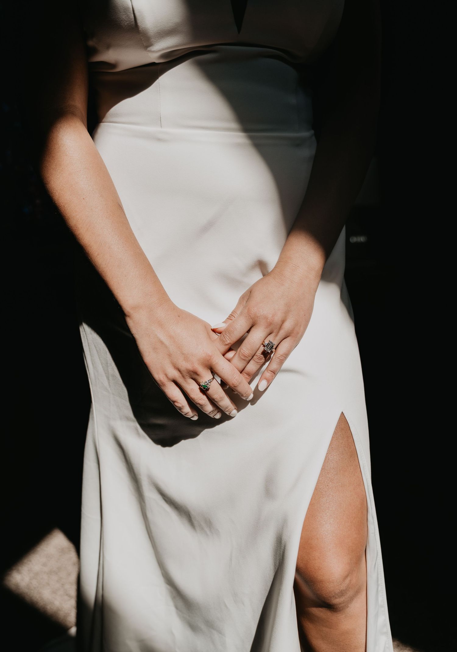 Close-up of hands with rings resting on a white dress with a side slit in dramatic lighting.