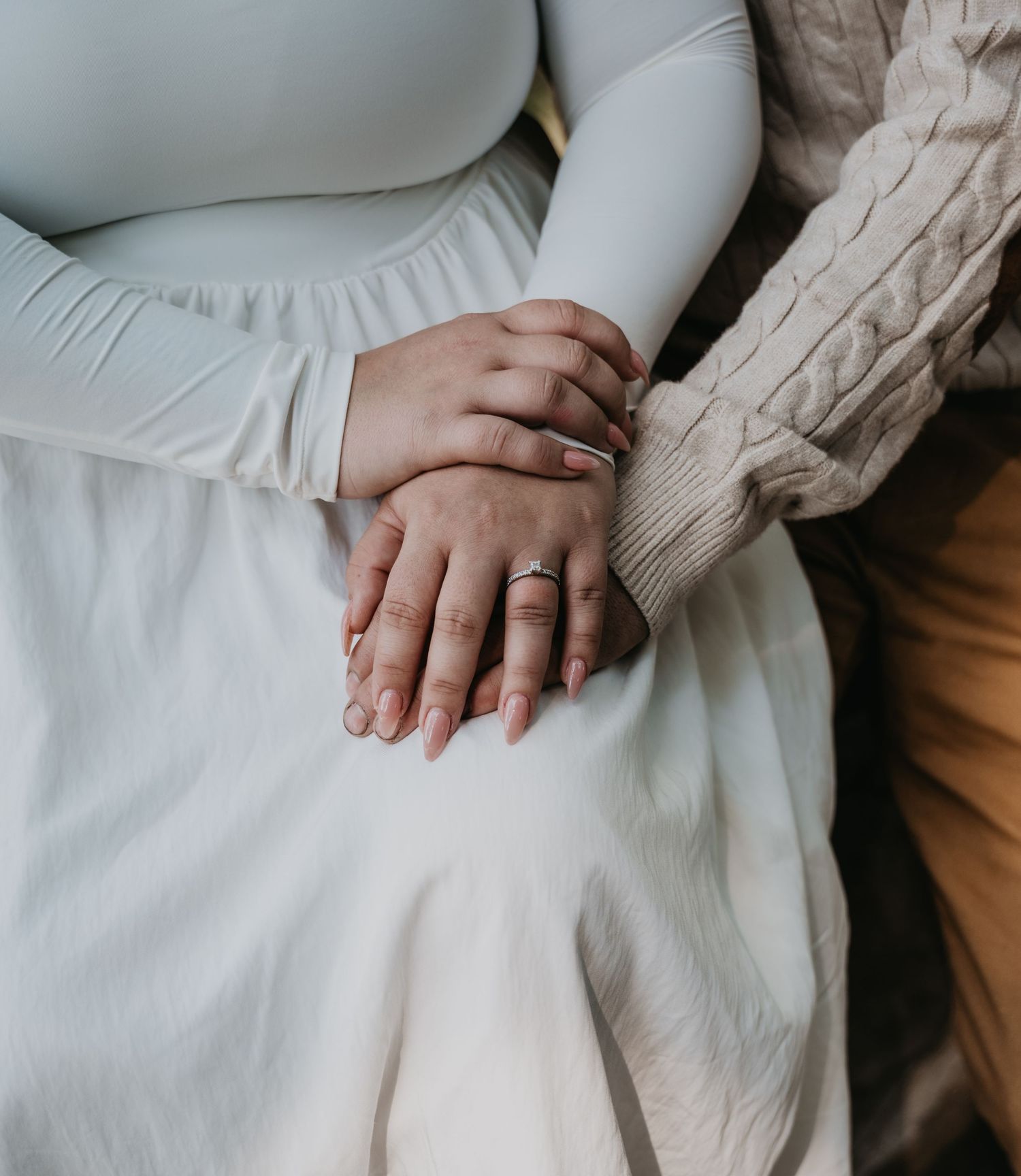 Two hands gently resting together on white fabric with a knit sweater sleeve visible in soft natural lighting.