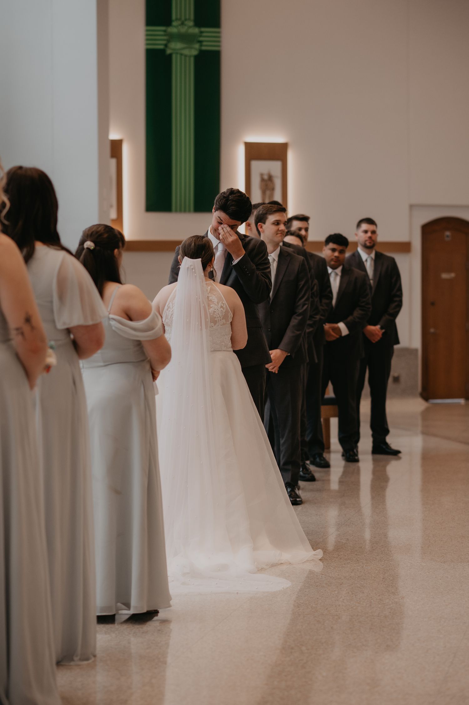 Wedding party gathers in a bright indoor venue with bridesmaids in gray dresses and groomsmen in dark suits.