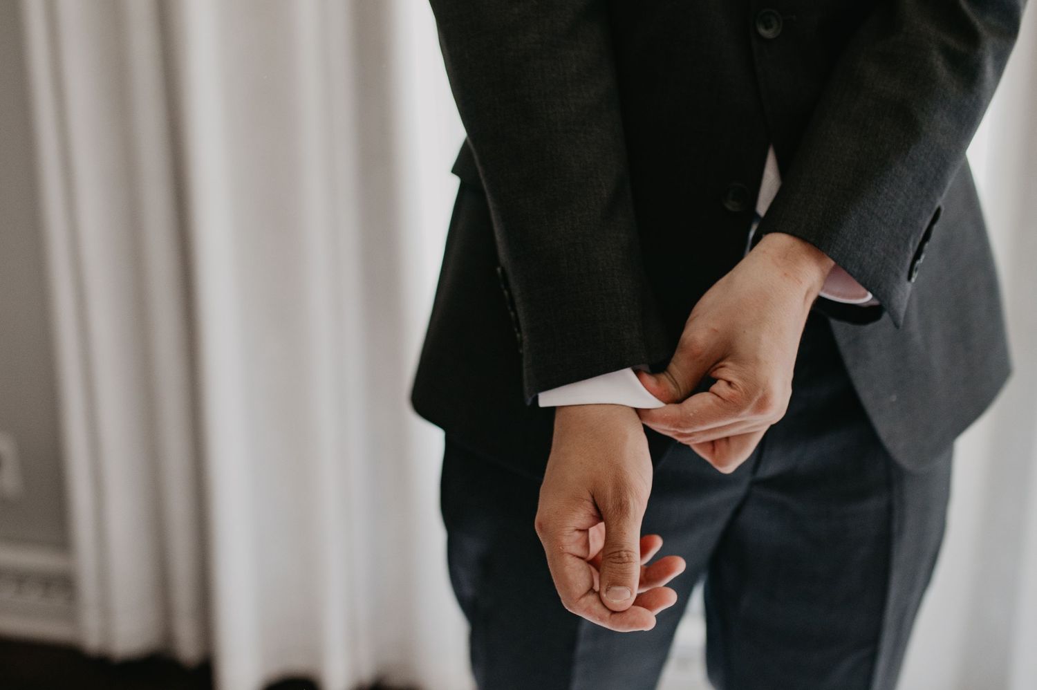 A person in a dark suit jacket adjusts their sleeve cuff while standing against a light colored background.