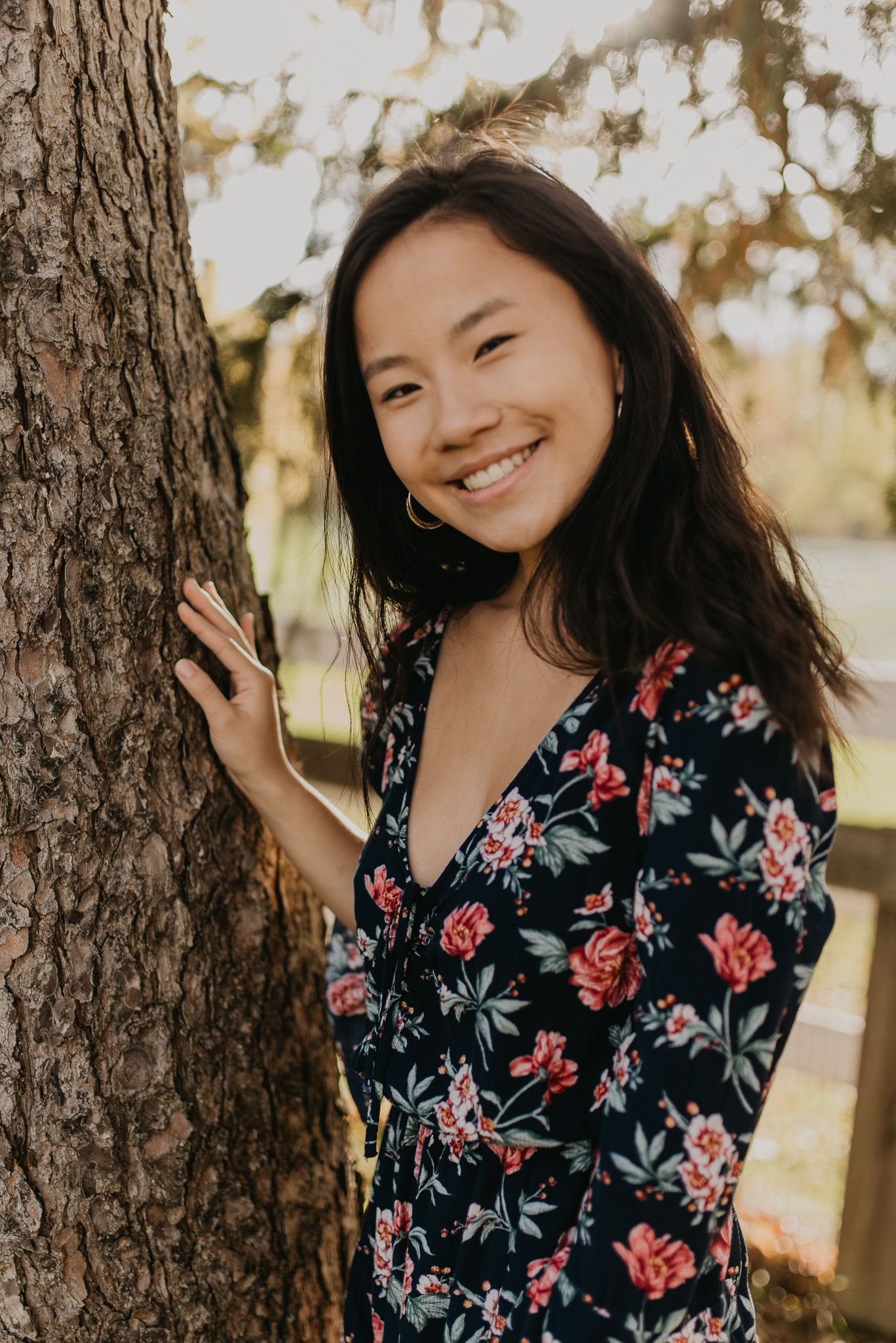 Someone wearing a floral print dress poses joyfully against a tree trunk in natural lighting.