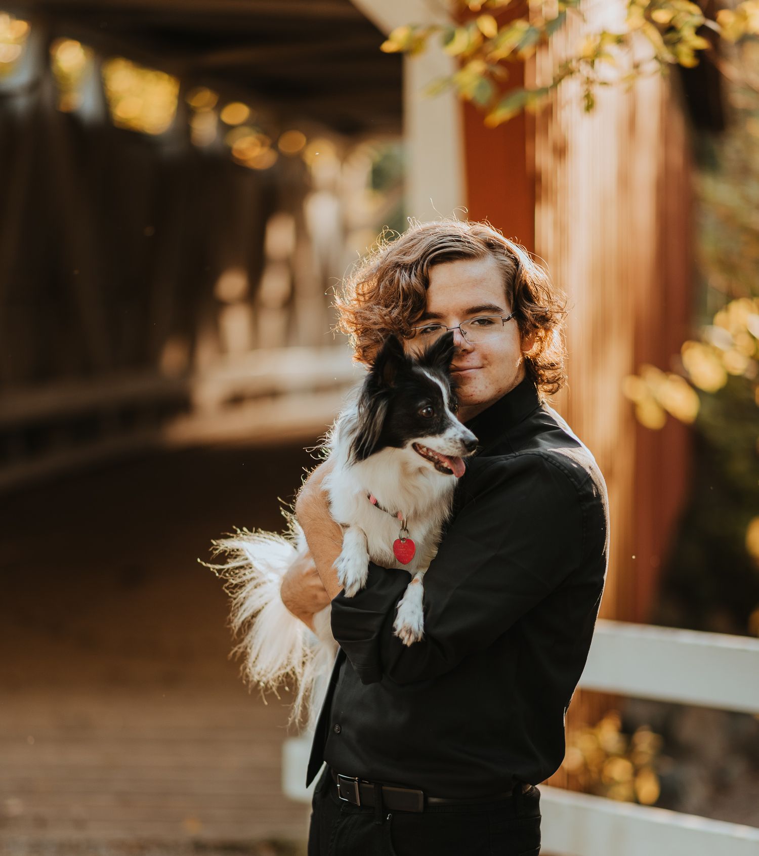 A young person in black attire holds and cuddles a happy white and fluffy Japanese Chin dog on a porch with bokeh lights.