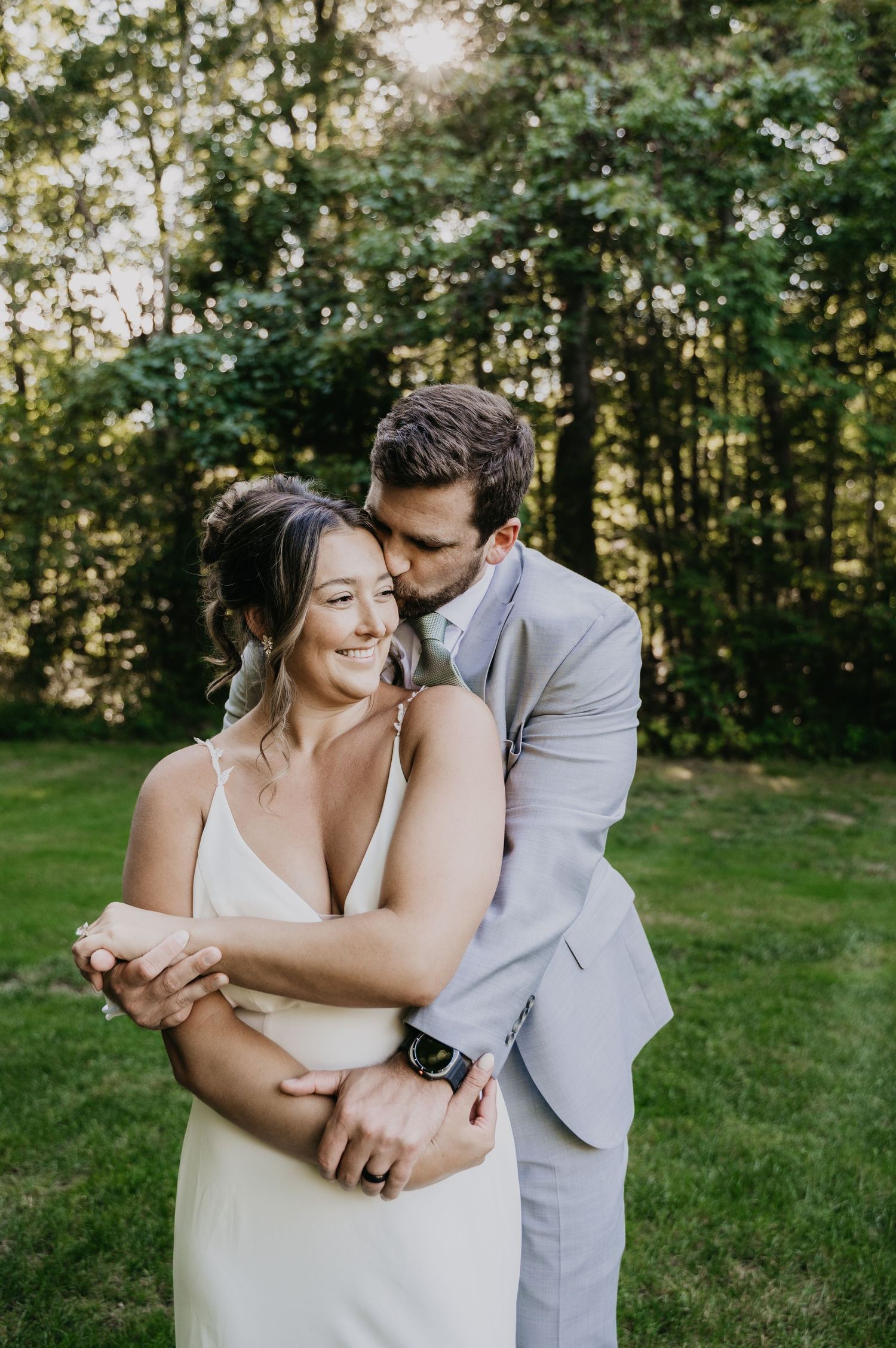 A romantic moment captured between newlyweds embracing on a sunlit lawn with trees in the background.