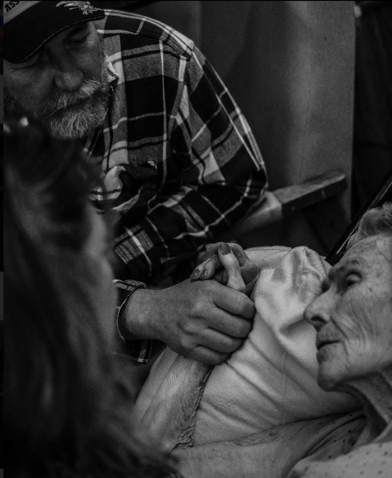 Black and white photo of an elderly person lying in bed holding hands with a bearded man in a plaid shirt.