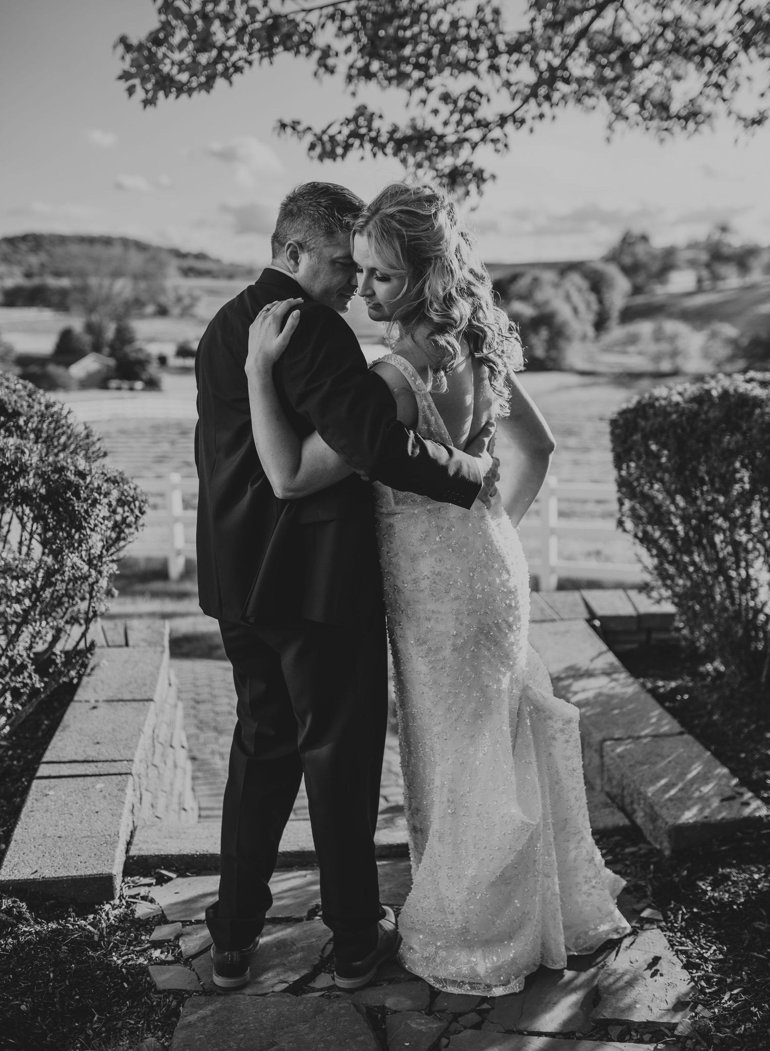 Black and white photo of newlyweds embracing with scenic countryside view.