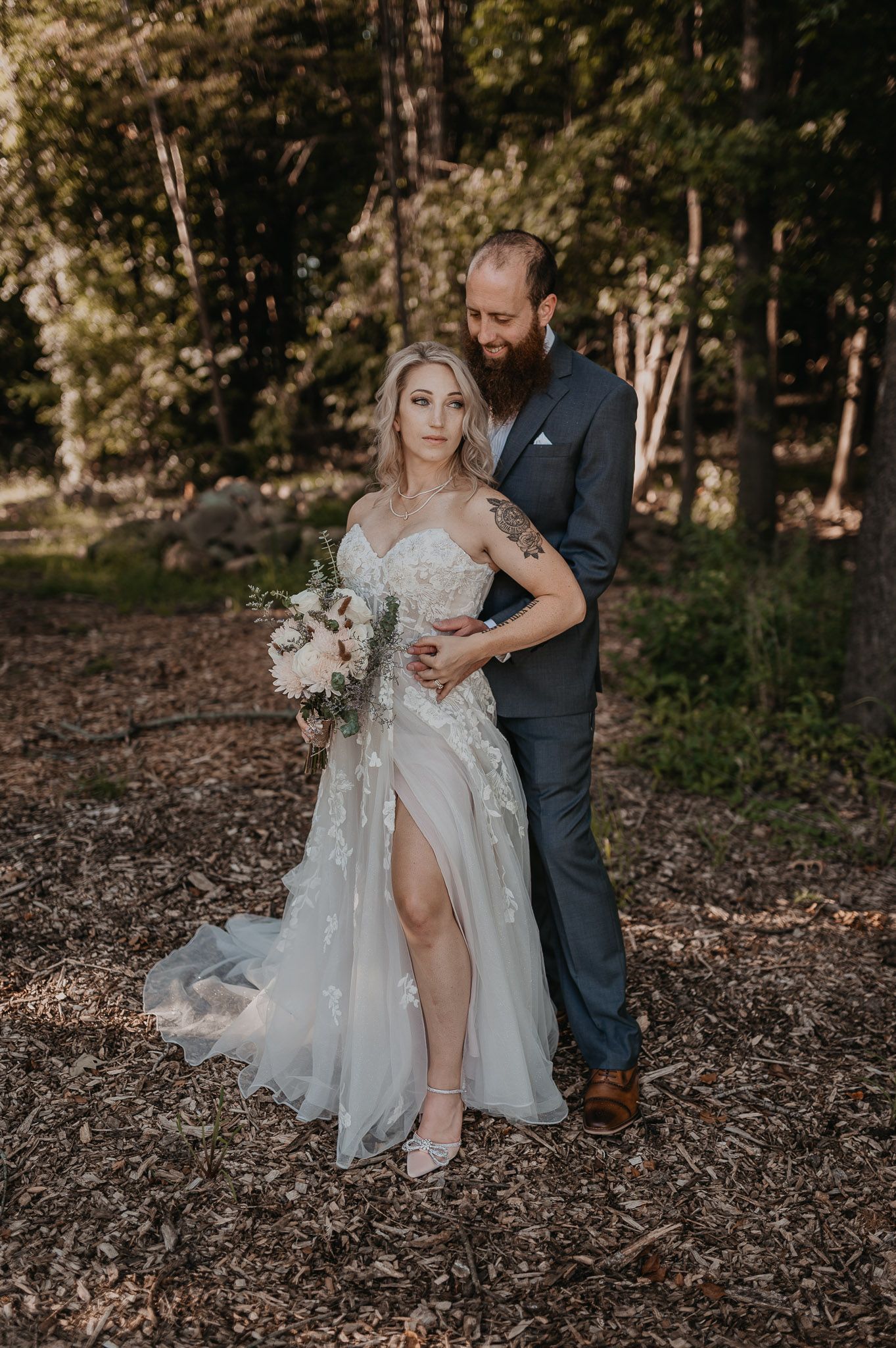 Bride in flowing lace gown with thigh slit poses with groom among autumn trees.