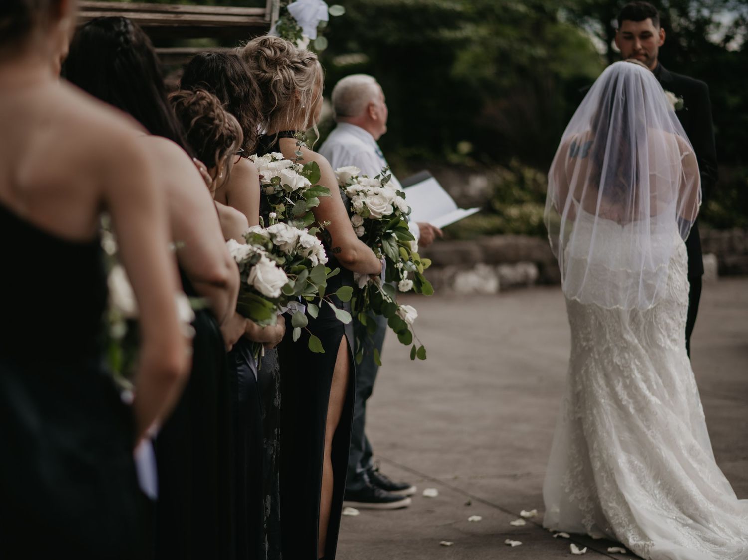 Wedding party stands in line holding elegant white floral bouquets as bride in lace gown and veil walks past.