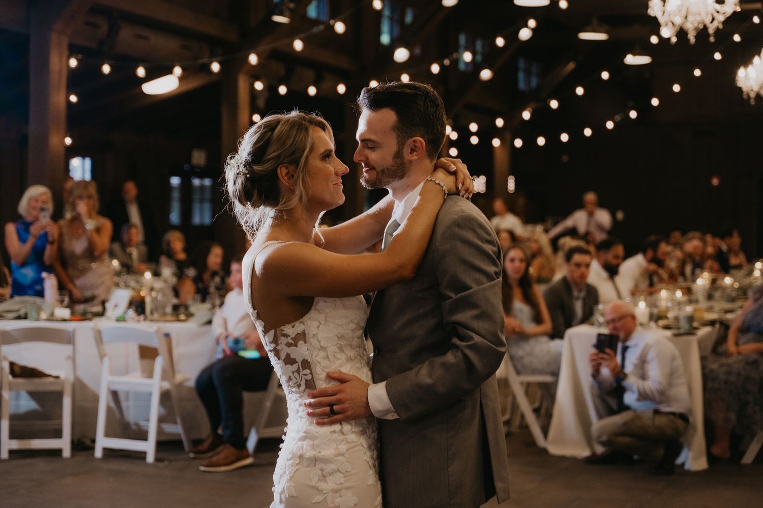 Newlyweds share their first dance under twinkling lights at rustic wedding reception.
