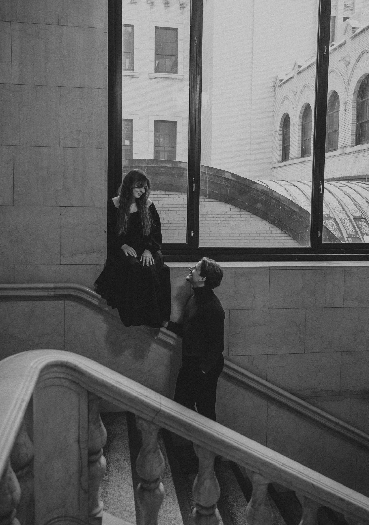 A black and white photograph showing two people having a conversation on an elegant curved staircase with stone balustrades.