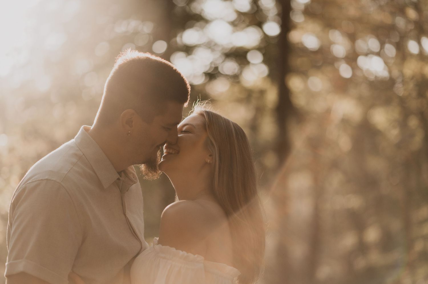 A romantic couple shares an intimate kiss in a golden-lit forest setting during a wedding photoshoot.