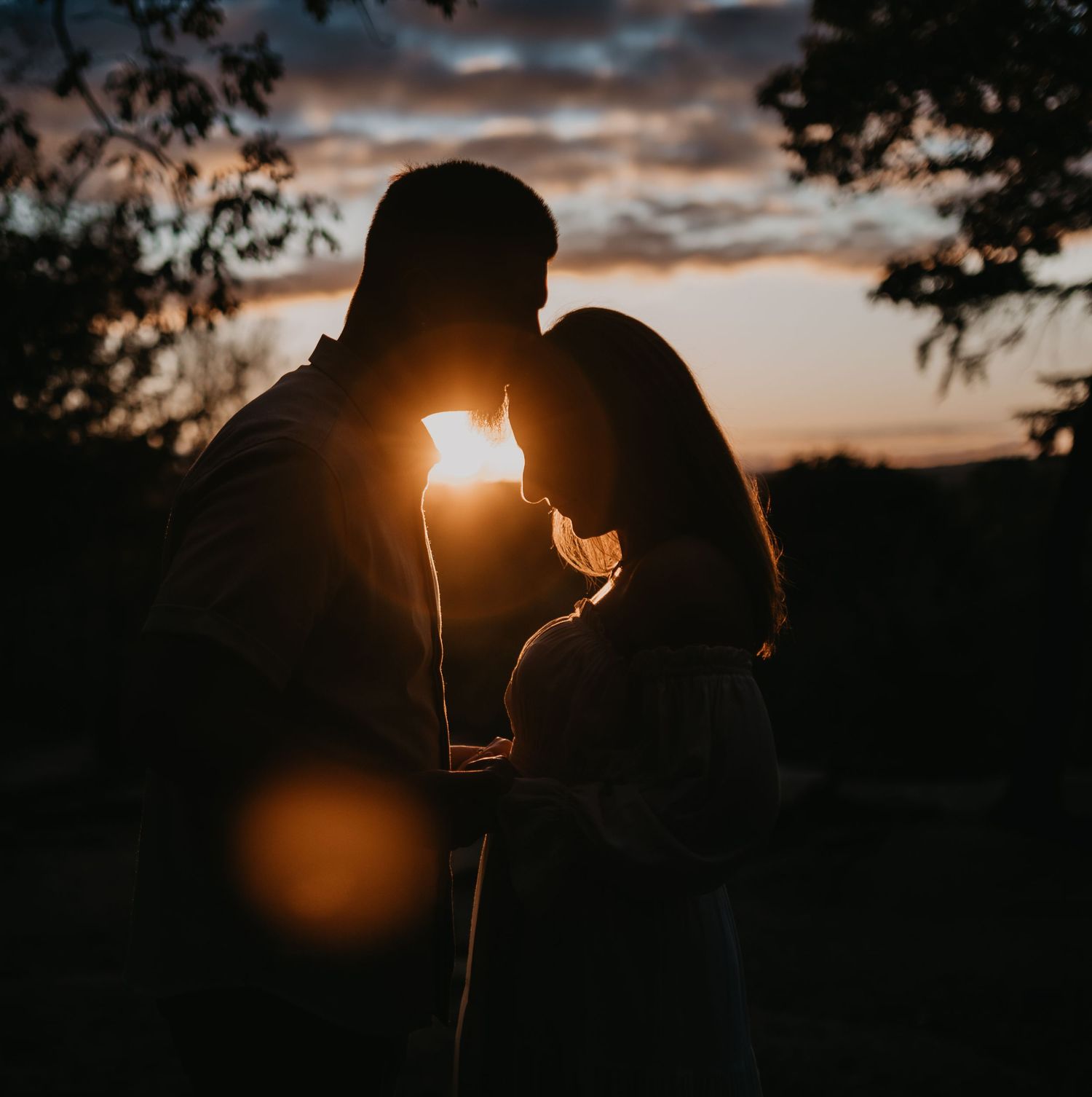 Silhouetted couple embracing against a dramatic sunset sky with golden light peeking through.