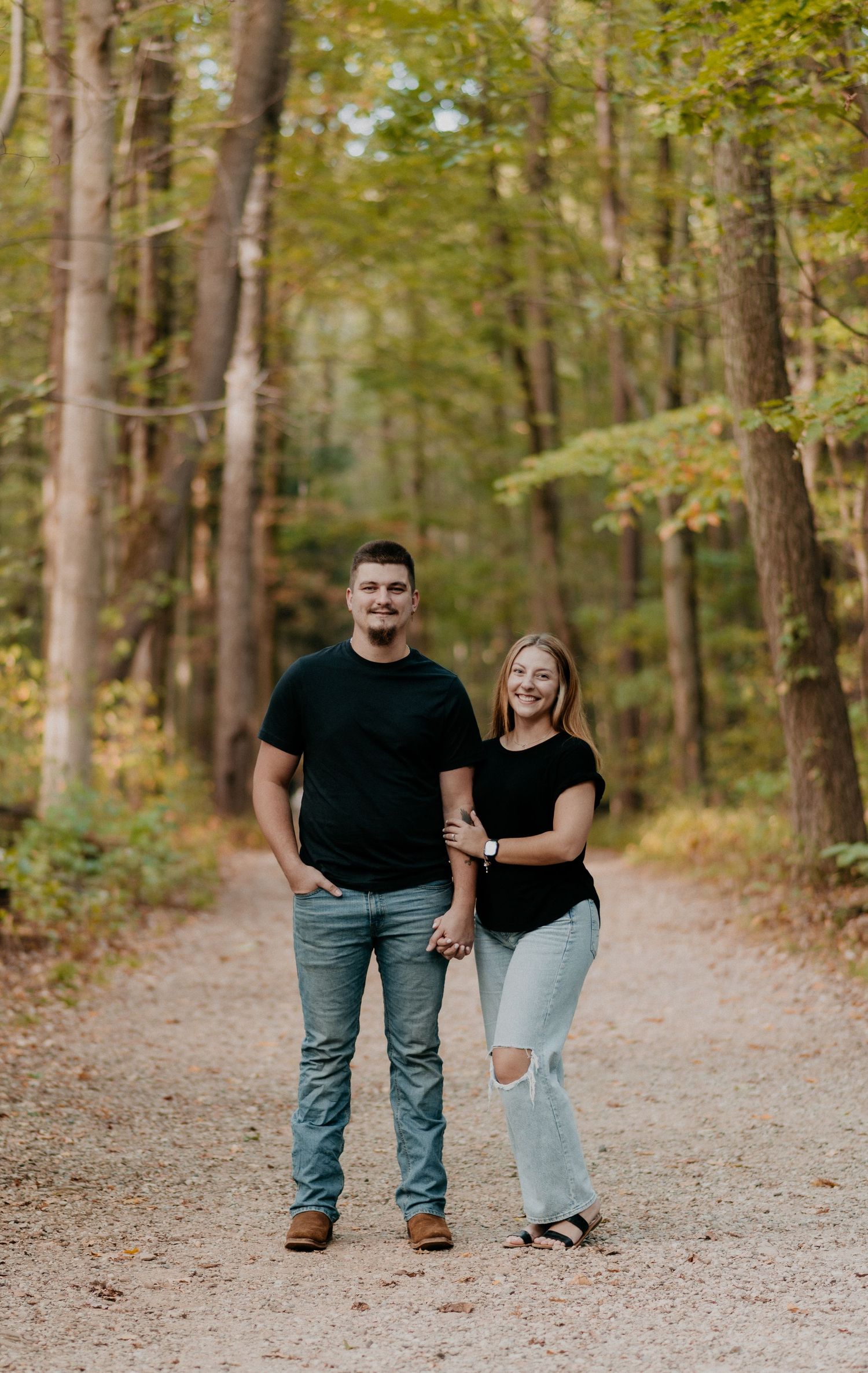 A couple walks together on a wooded trail surrounded by tall trees on an autumn afternoon.