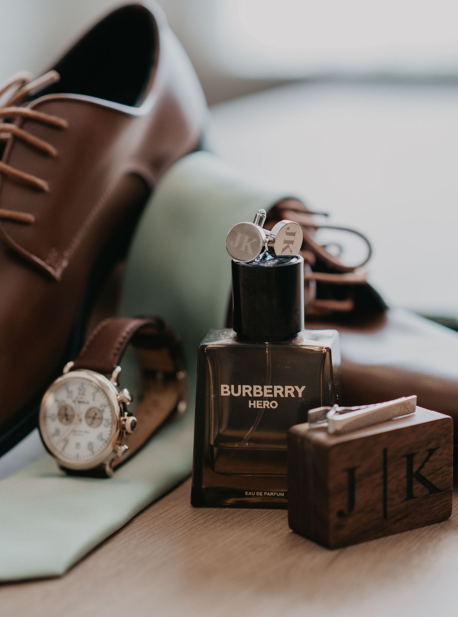 Wedding accessories including brown leather shoes, watch, Burberry cologne and wooden box arranged on light surface.