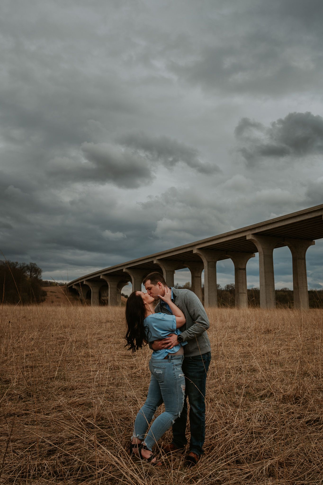 A couple shares a tender moment in a field with a dramatic bridge backdrop and stormy sky.