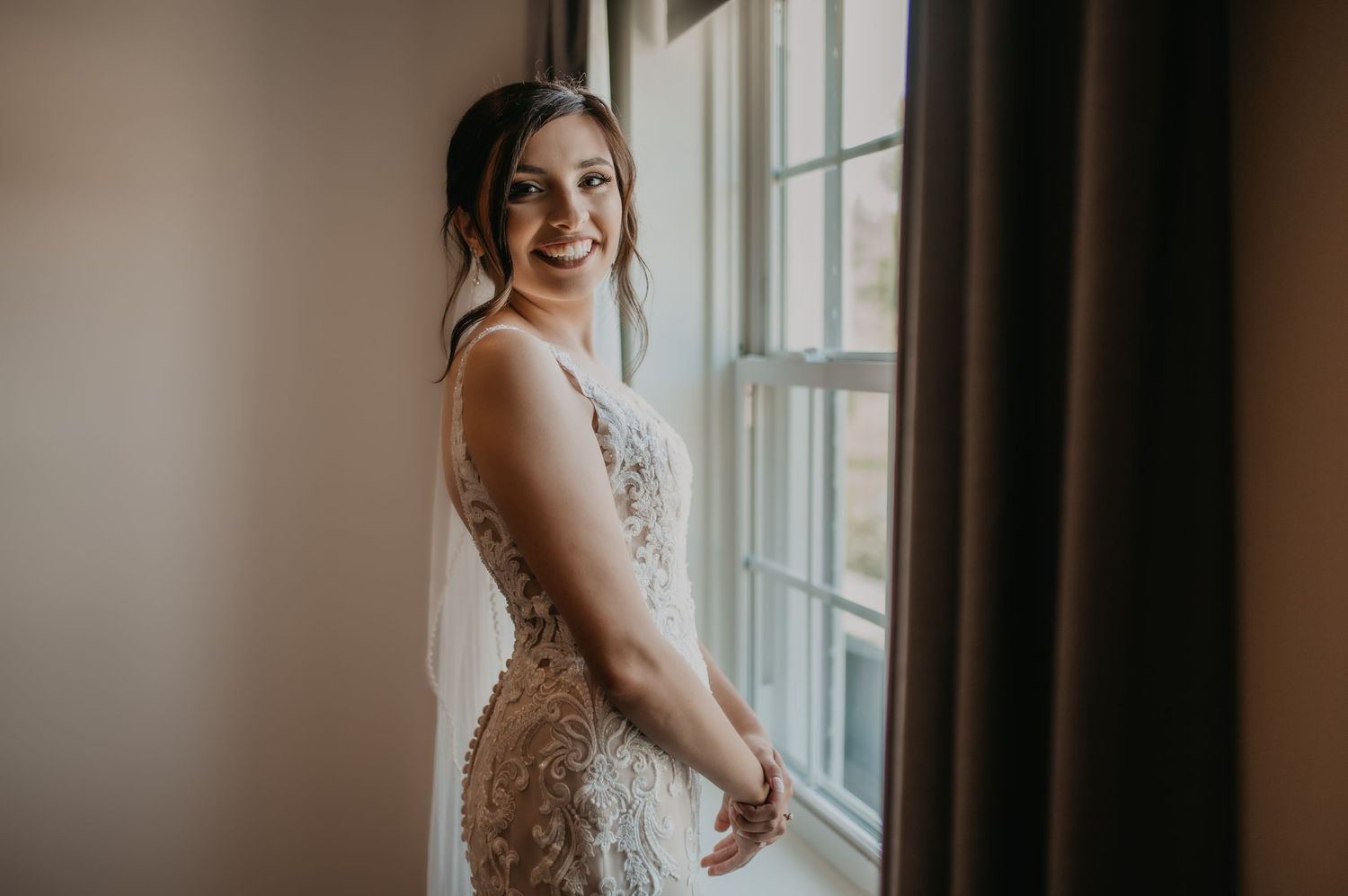 Elegant bride in lace dress posing by window in soft natural light.
