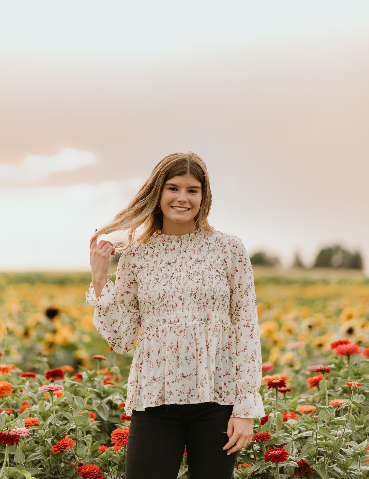 A woman stands in a colorful field of sunflowers and red poppies wearing a floral long-sleeved blouse.