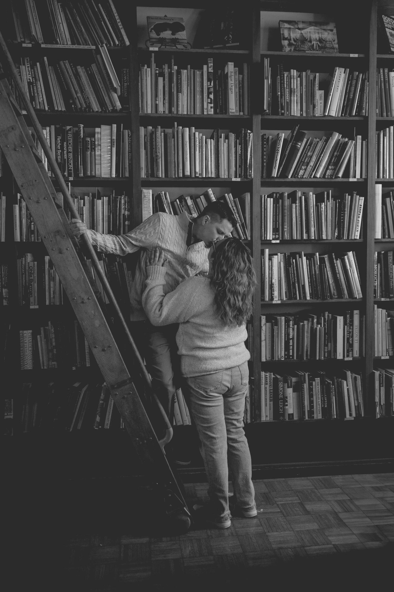 A romantic moment between two people sharing a kiss on a library ladder surrounded by towering bookshelves.